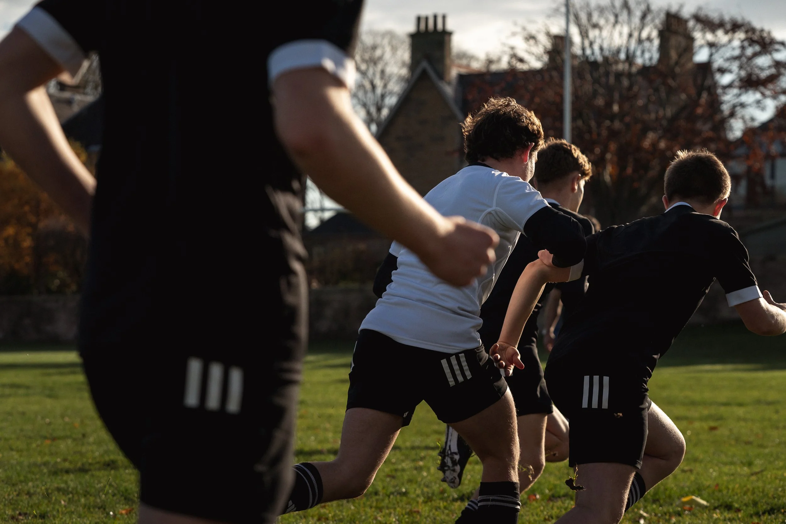 A group of young male rugby players in Adidas kit running in a line on a grassy field during late afternoon or early evening, with trees and houses in the background.