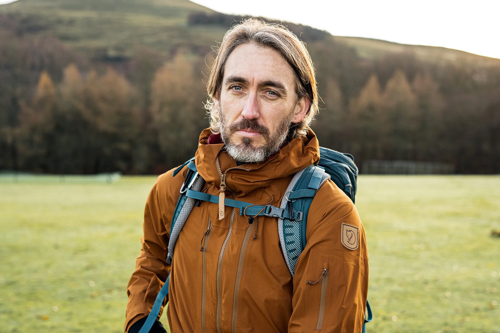 A man with gray hair and beard, wearing an orange jacket and hiking gear, standing outdoors on a grassy area with hills and trees in the background.
