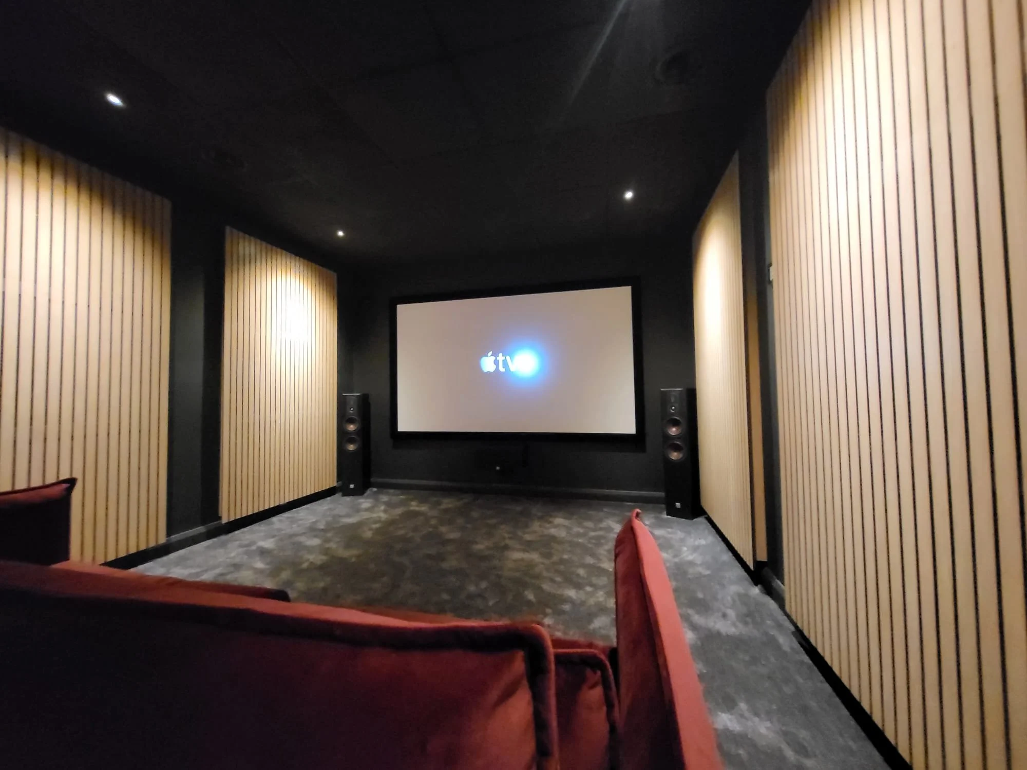 Acoustic Panels in A home movie theater room with red seats, wooden panel walls, dark ceiling with small lights, and a large screen displaying the Apple TV logo.