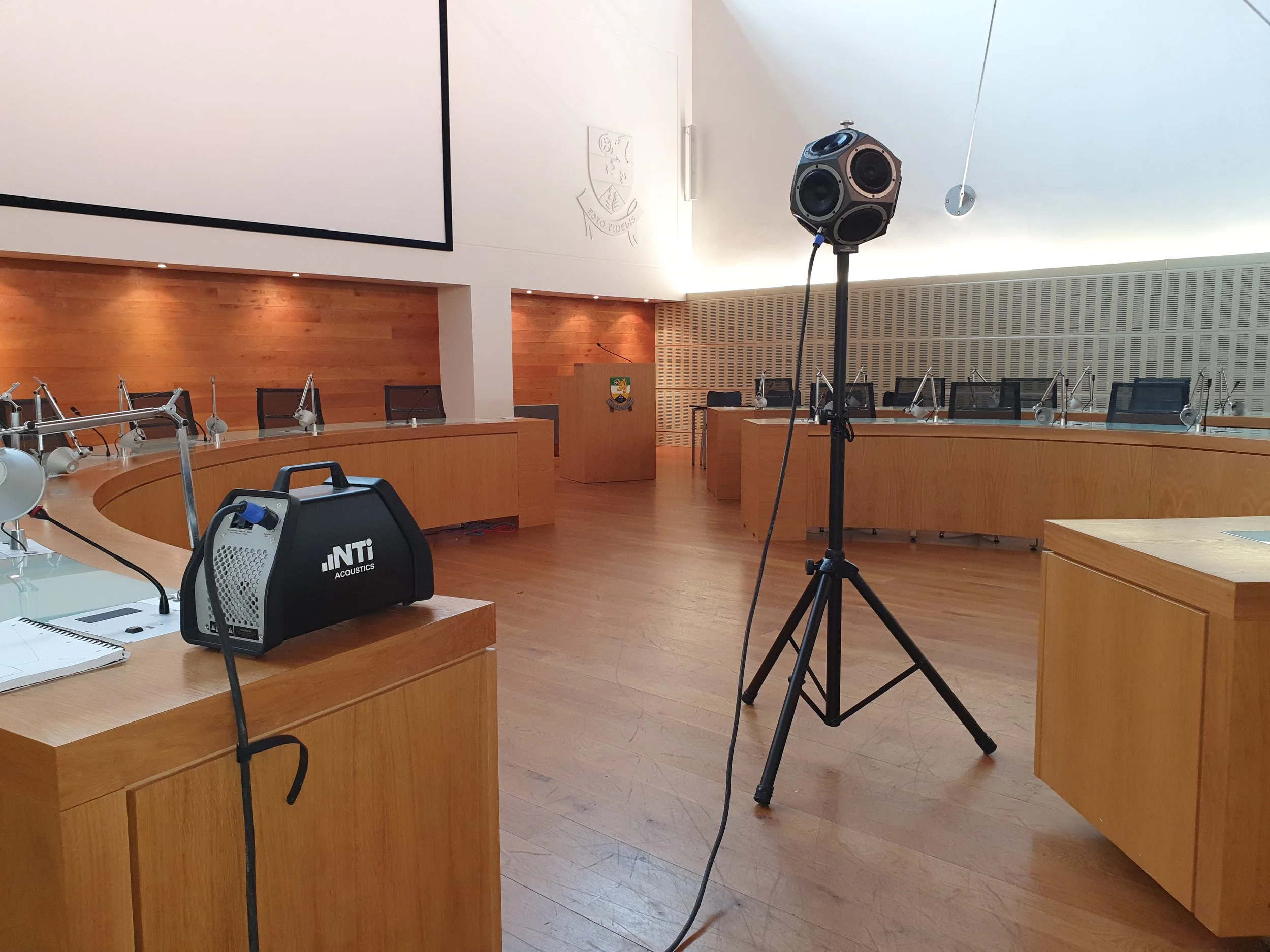 Acoustic Testing in a conference or meeting room with wooden tables and chairs, a large speaker on a stand, a podium, and a large projection screen on the wall.