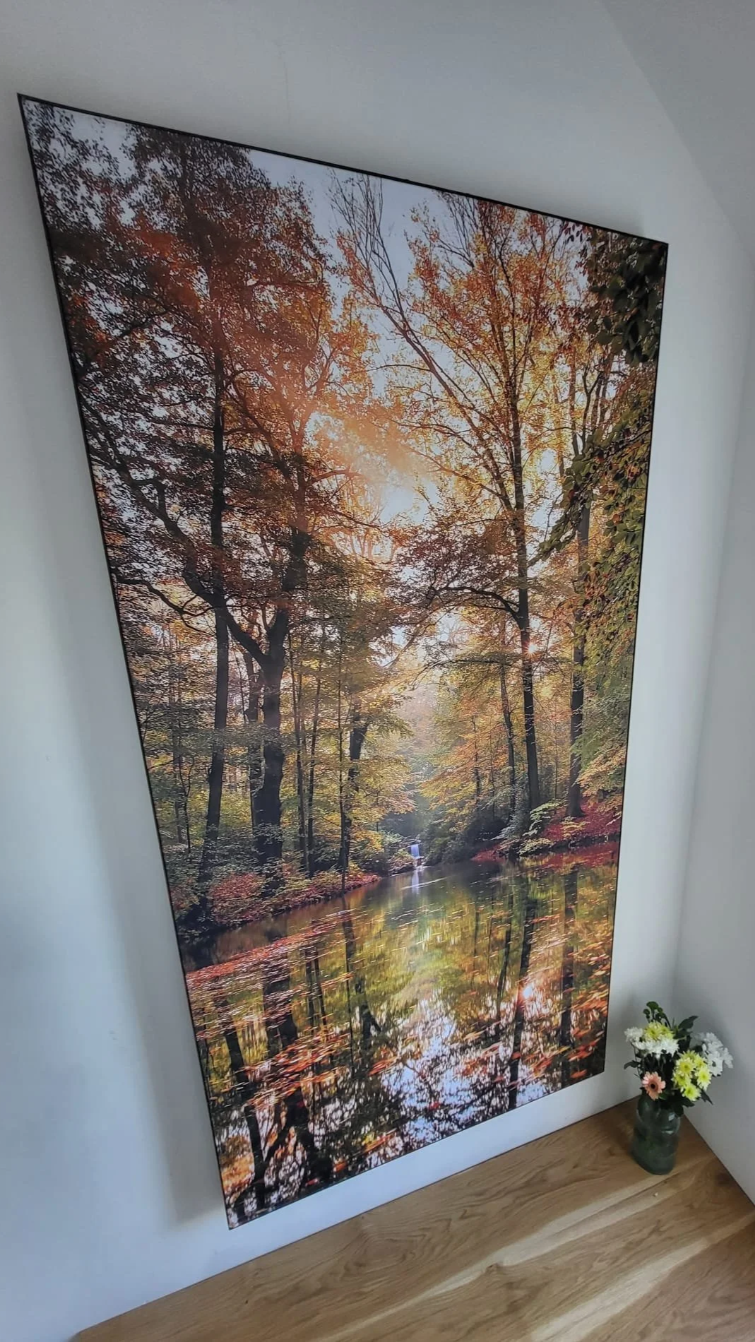 Framed artwork acoustic panel of an autumn forest with colorful fall trees and a reflection in a river on a white wall, with a small vase of flowers on the floor.