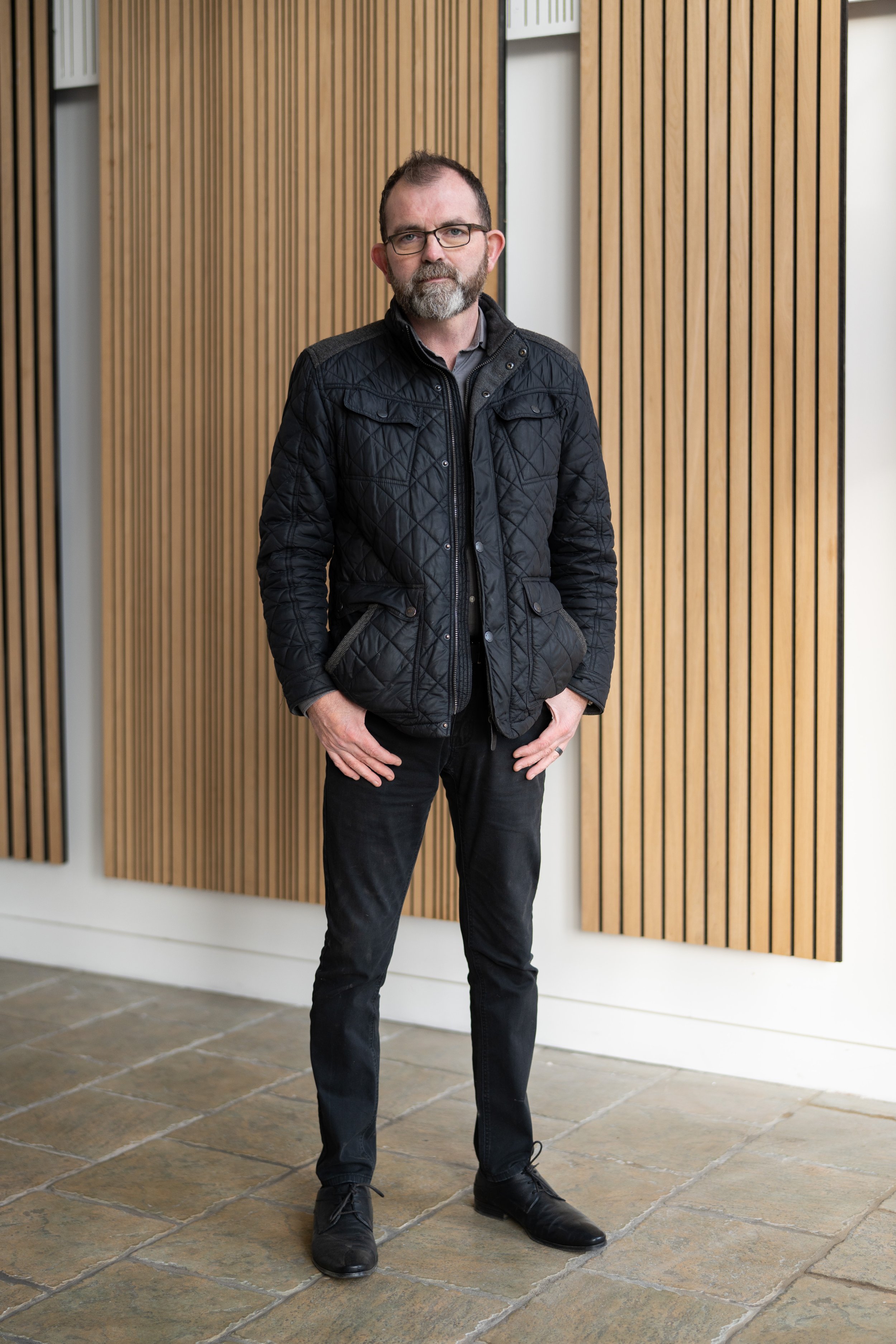 A man with glasses, short dark hair, and a beard standing indoors against a background of vertical wooden panels. He is wearing a black quilted jacket, black pants, and black shoes, with one hand in his pocket.