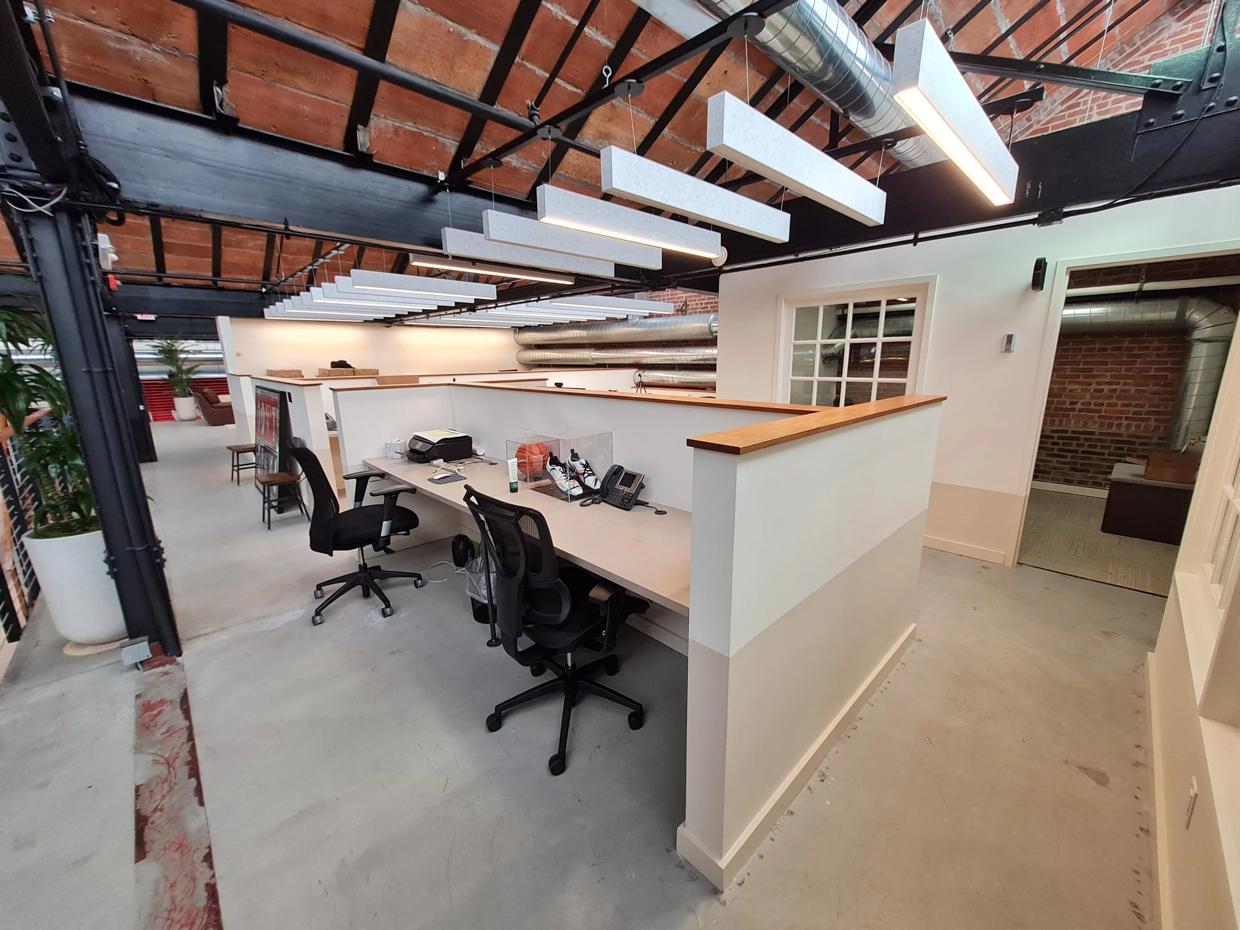 Acoustic Ceiling Baffles in a modern Office space with a white reception desk, black chairs, a computer, and a shoe display case. Overhead hanging lights and exposed ductwork on ceiling with brick walls.