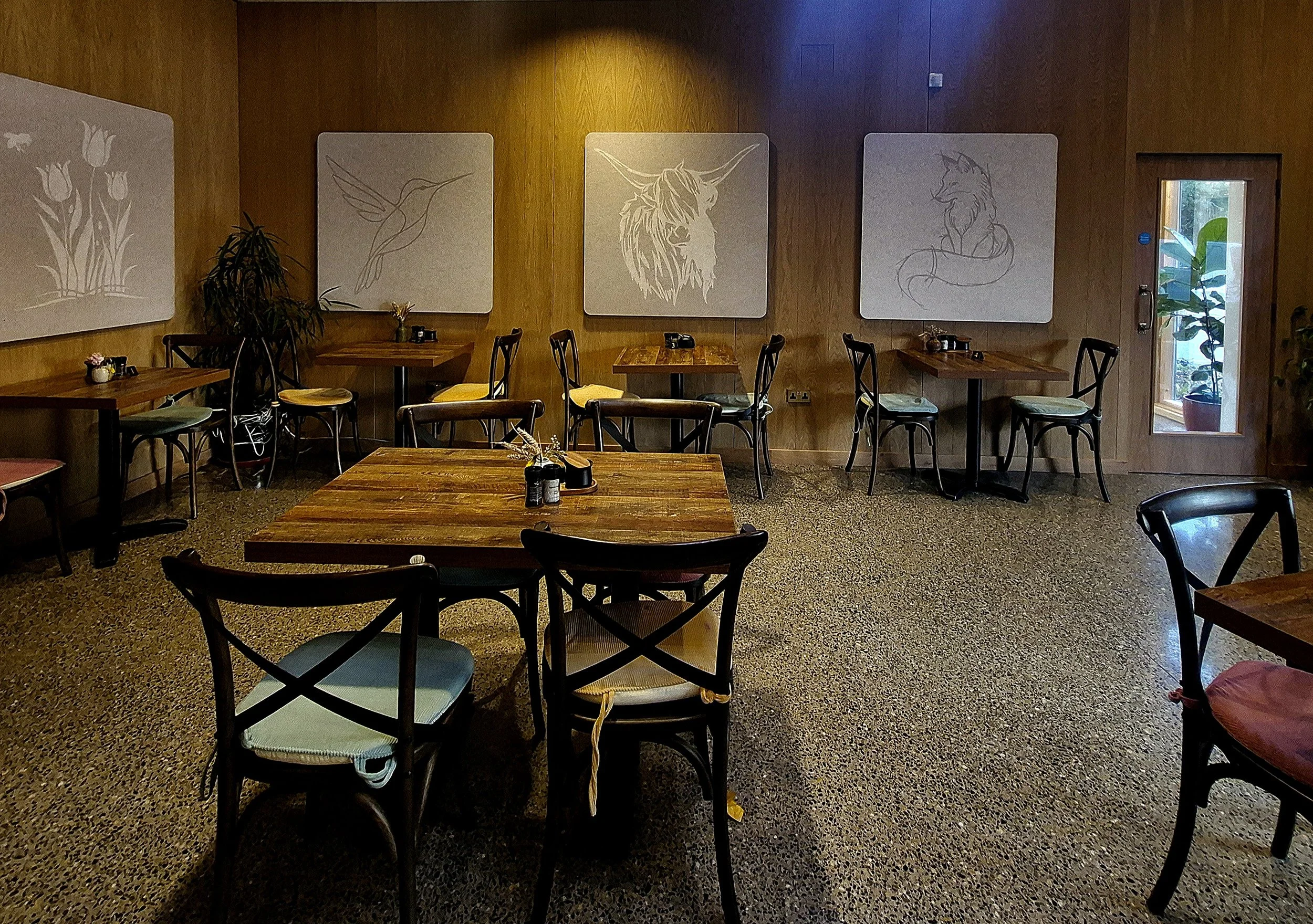 Bespoke PET acoustic wall panels Interior of a restaurant with wooden tables and chairs, large art panels on the wall featuring abstract line drawings of animals, a plant near the wall, and a glass door with a plant outside visible.