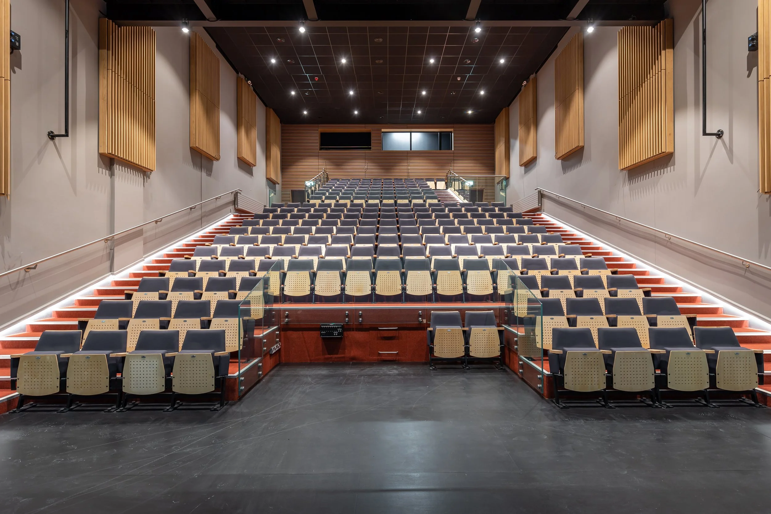 Acoustic treatment in an empty auditorium with tiered seating, black flooring, and wooden wall panels. Small black speakers are mounted on the side walls, and the ceiling has multiple recessed lights.