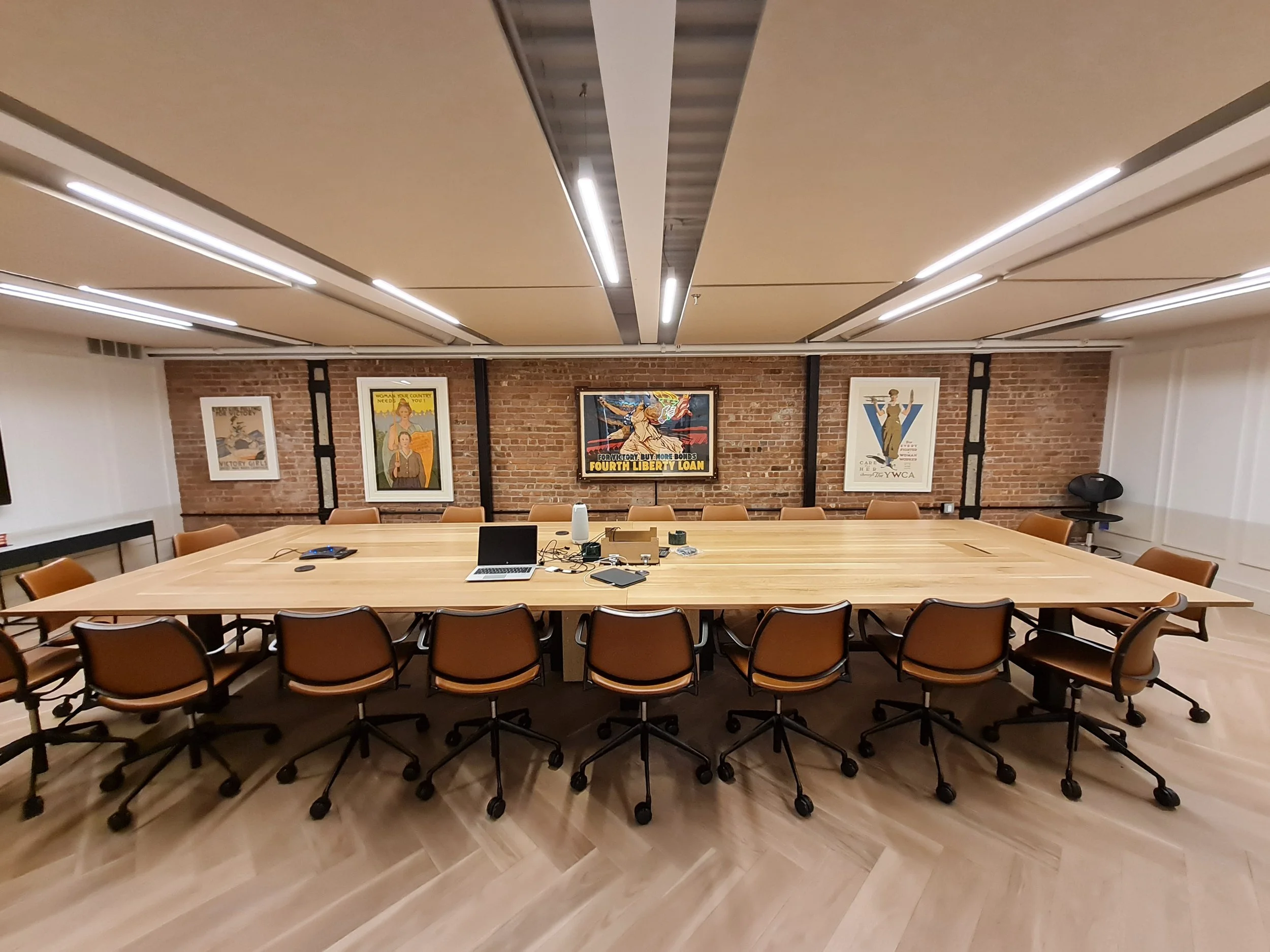 A conference room with a large wooden table surrounded by brown chairs, with a brick wall decorated with framed vintage posters, a laptop, and electronic devices on the table, and an acoustically treeated ceiling with modern lighting.