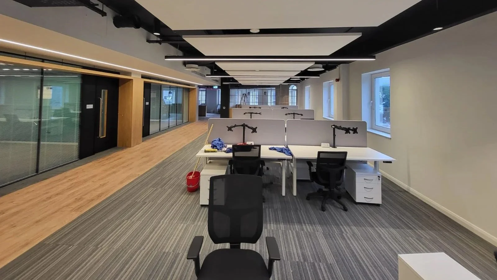 Acoustic Ceiling Panels in Empty office space with desks, chairs, and partition screens, with some cleaning cloths and a mop on the floor, windows providing natural light.