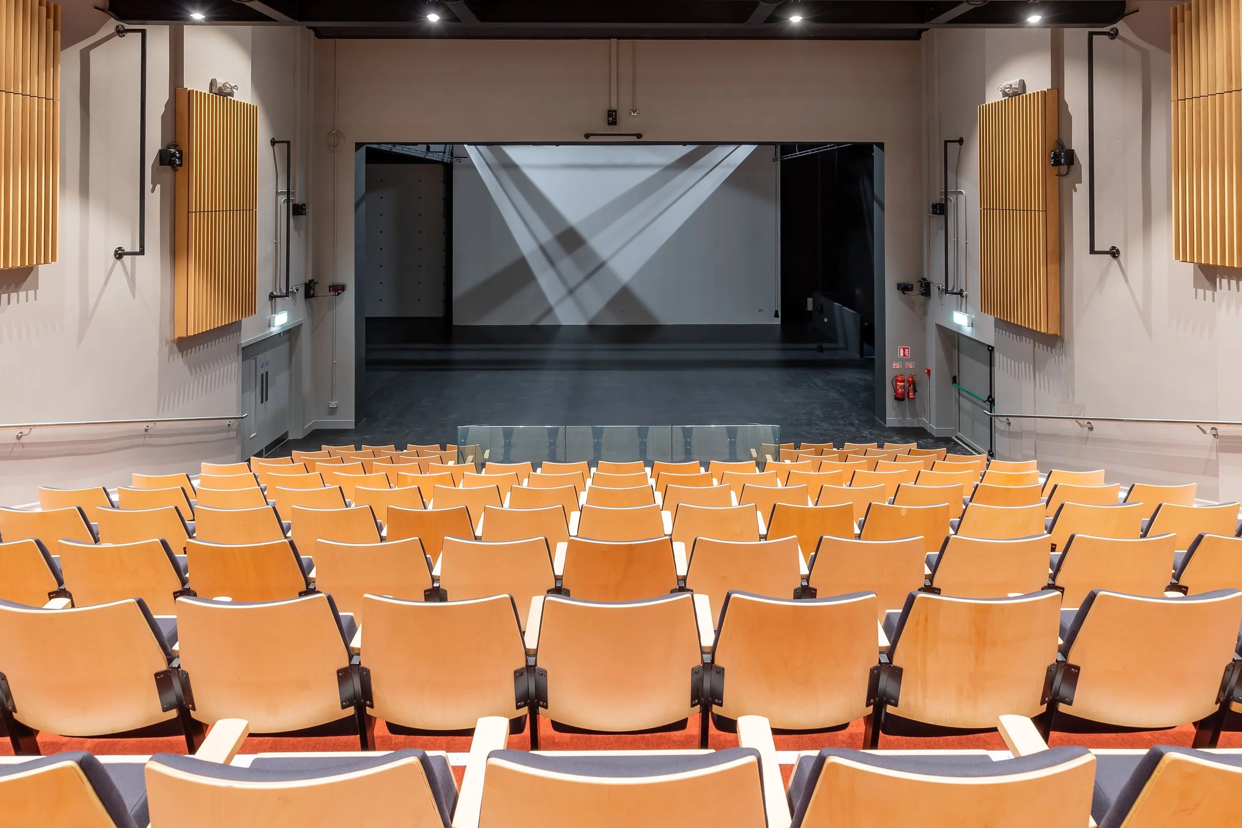 An auditorium or theater with a stage, rows of orange chairs, wood paneling on the walls, black ceiling, and spotlights, ready for a performance or presentation.