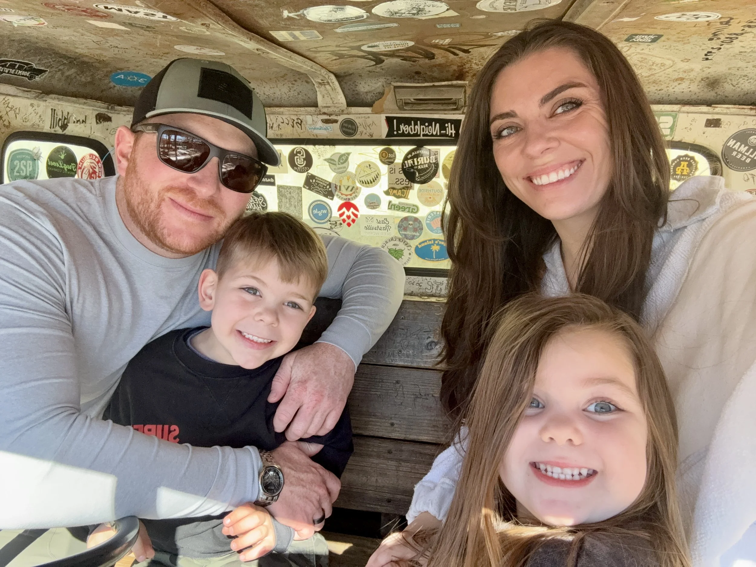 Family of four taking a selfie inside a vehicle covered in stickers, with two children in the front, a woman with long hair and a man wearing sunglasses and a cap in the back.