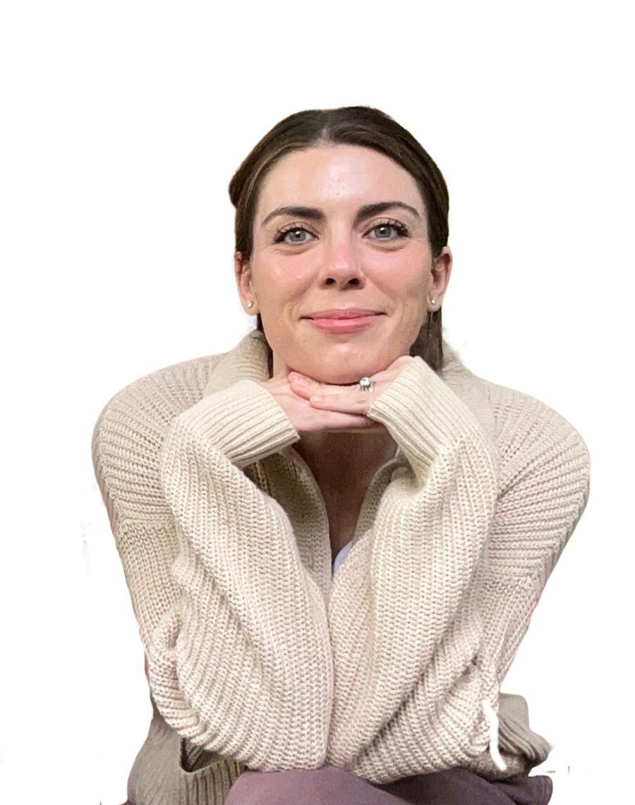 A woman with dark brown hair and light skin, smiling, sitting outdoors behind a wooden fence. She is resting her chin on her hands and wearing a beige knitted sweater.
