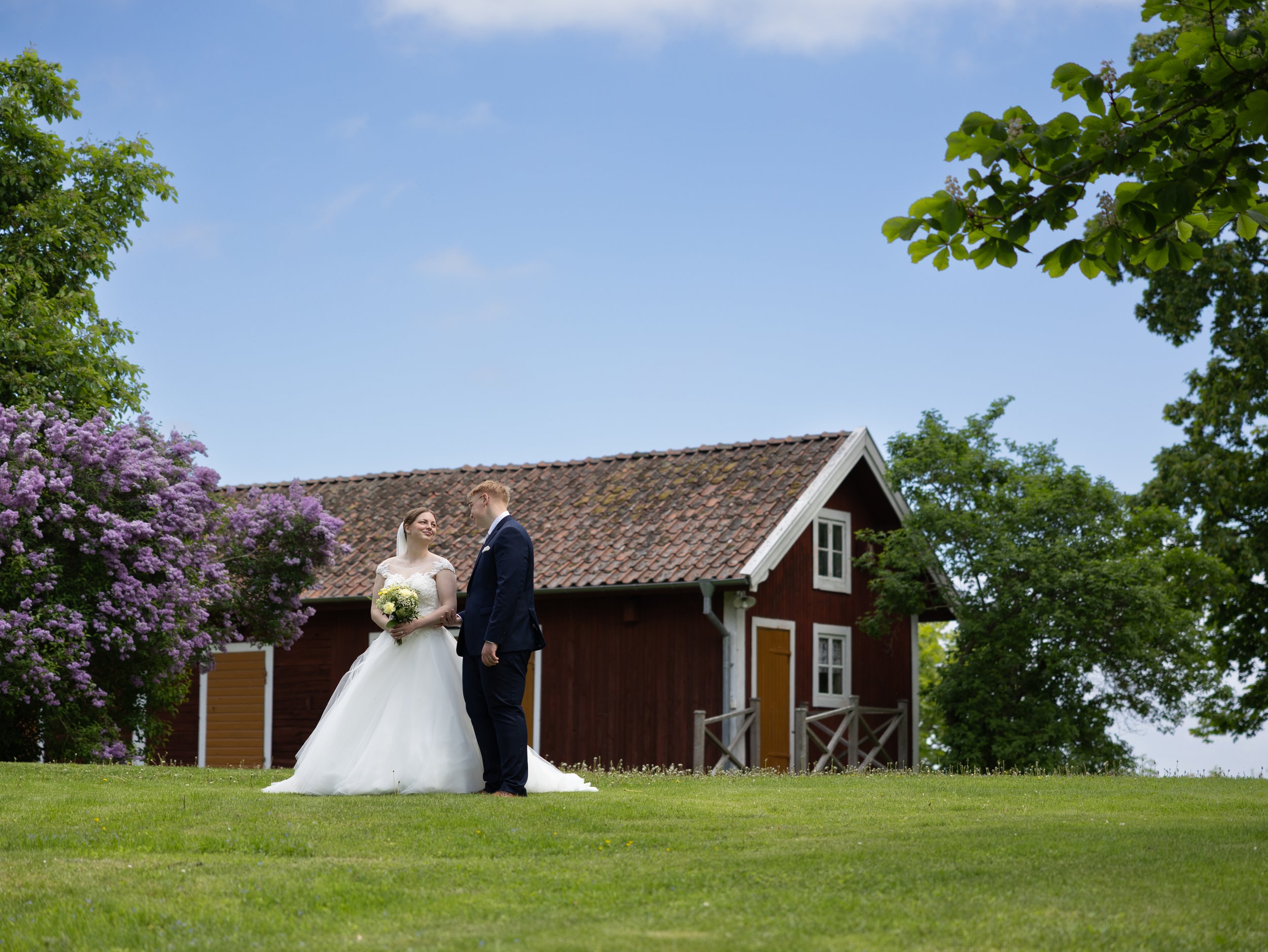 En brud och en brudgum står tillsammans utomhus framför ett rött hus med gröna träd och blommande buskar, under en klar blå himmel.