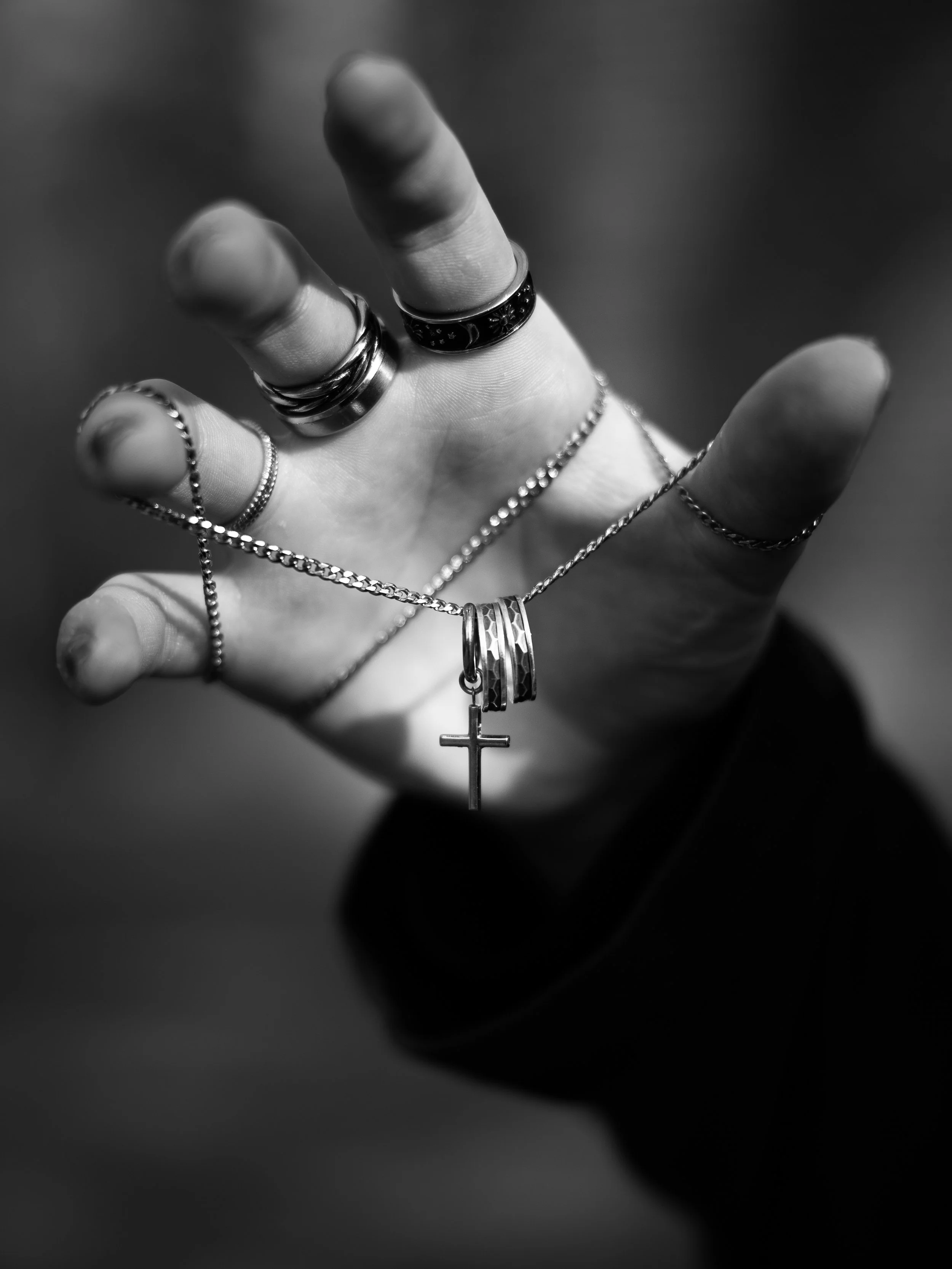 Svart och vitt fotografi av en hand som bär flera ringar och halsband med ett korshalsband, handens fingrar är utsträckta och fokuset är på ringarna och smyckena.