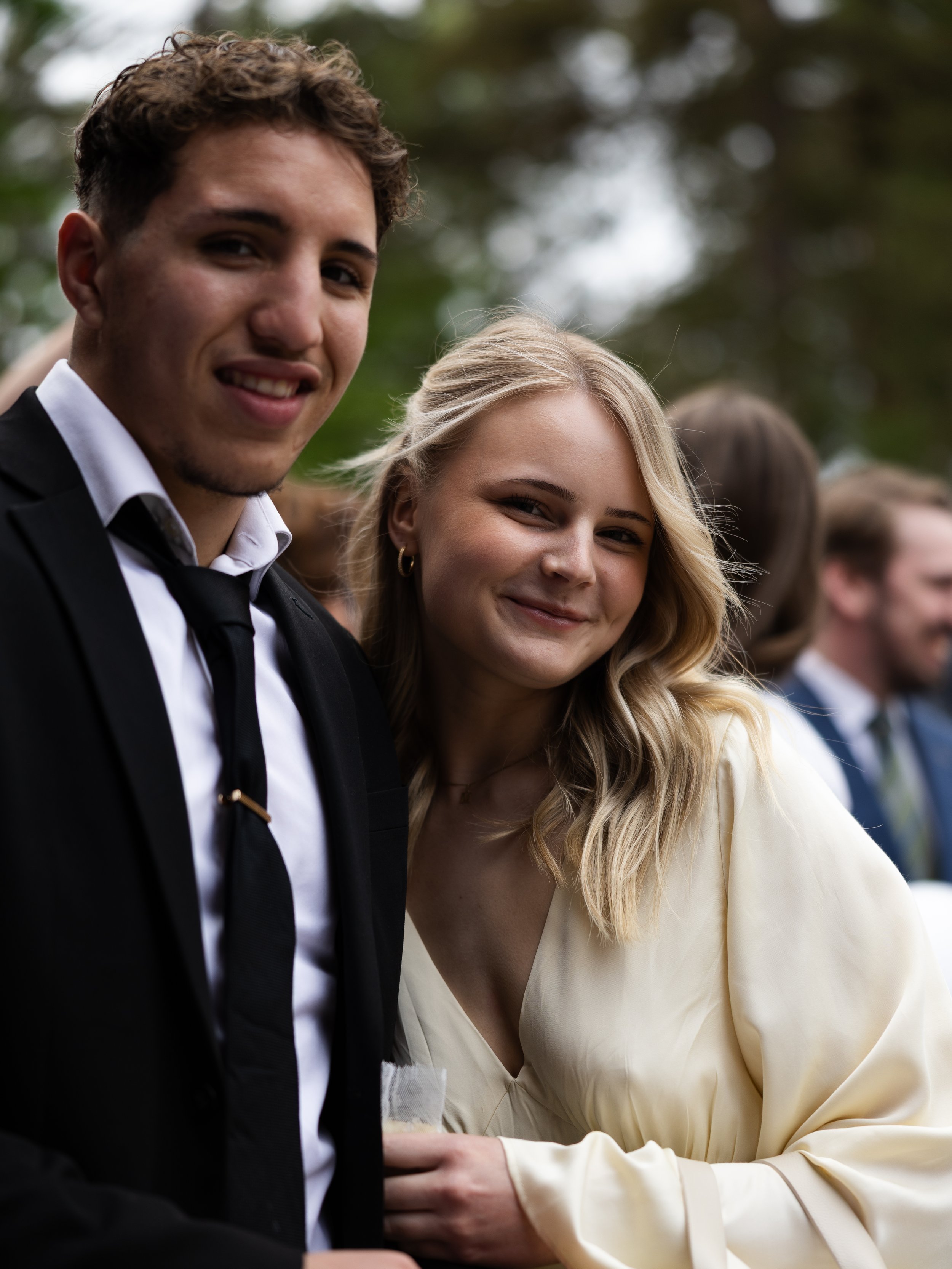Två unga vuxna personer, en man i svart kostym och vit skjorta och en kvinna i en ljus gul klänning, står nära varandra och ler utomhus.