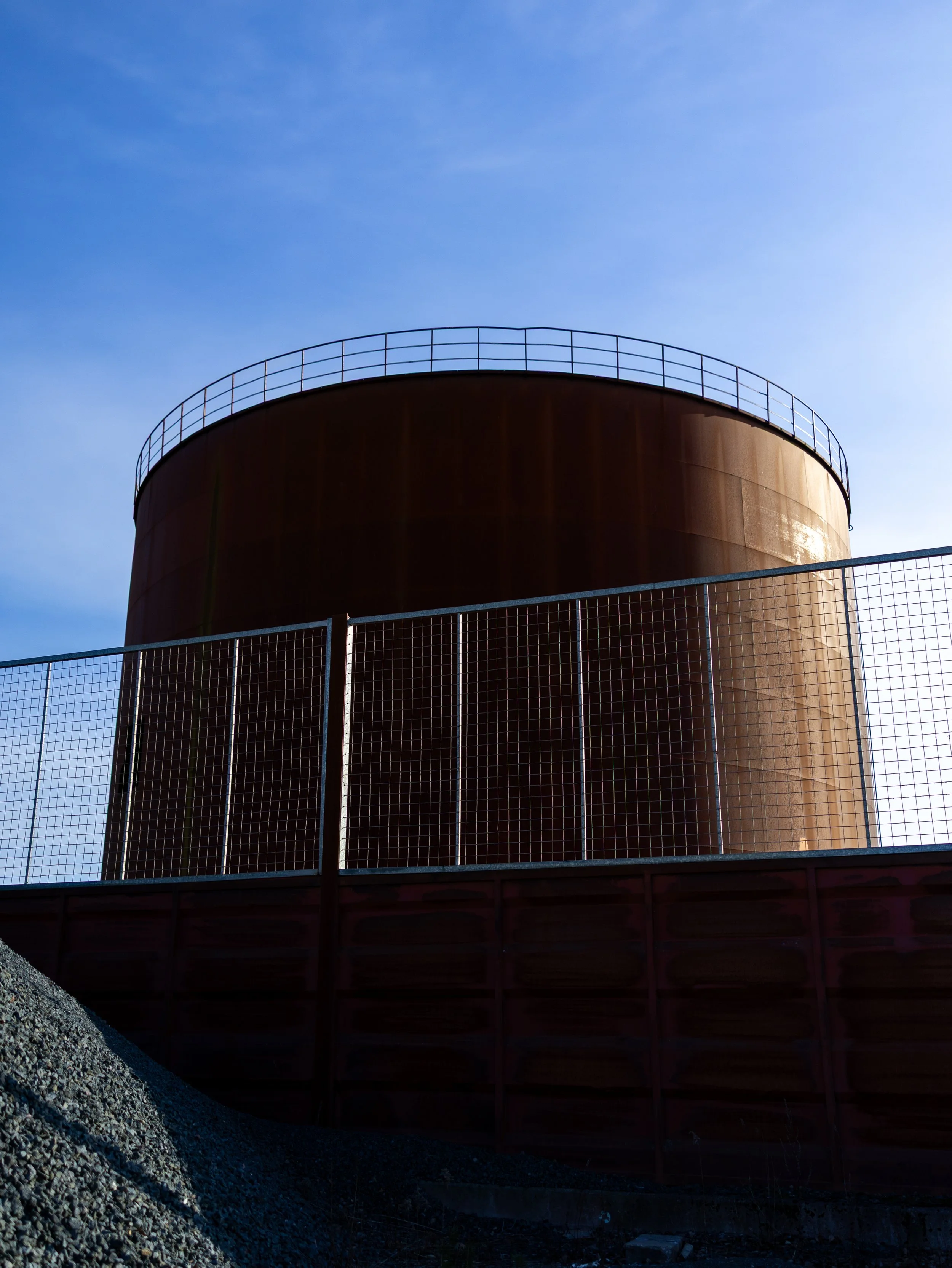 En stor, rund, rostfärgad industriell silos eller tank med ett säkerhetsräcke runt toppen, mot en klar blå himmel.