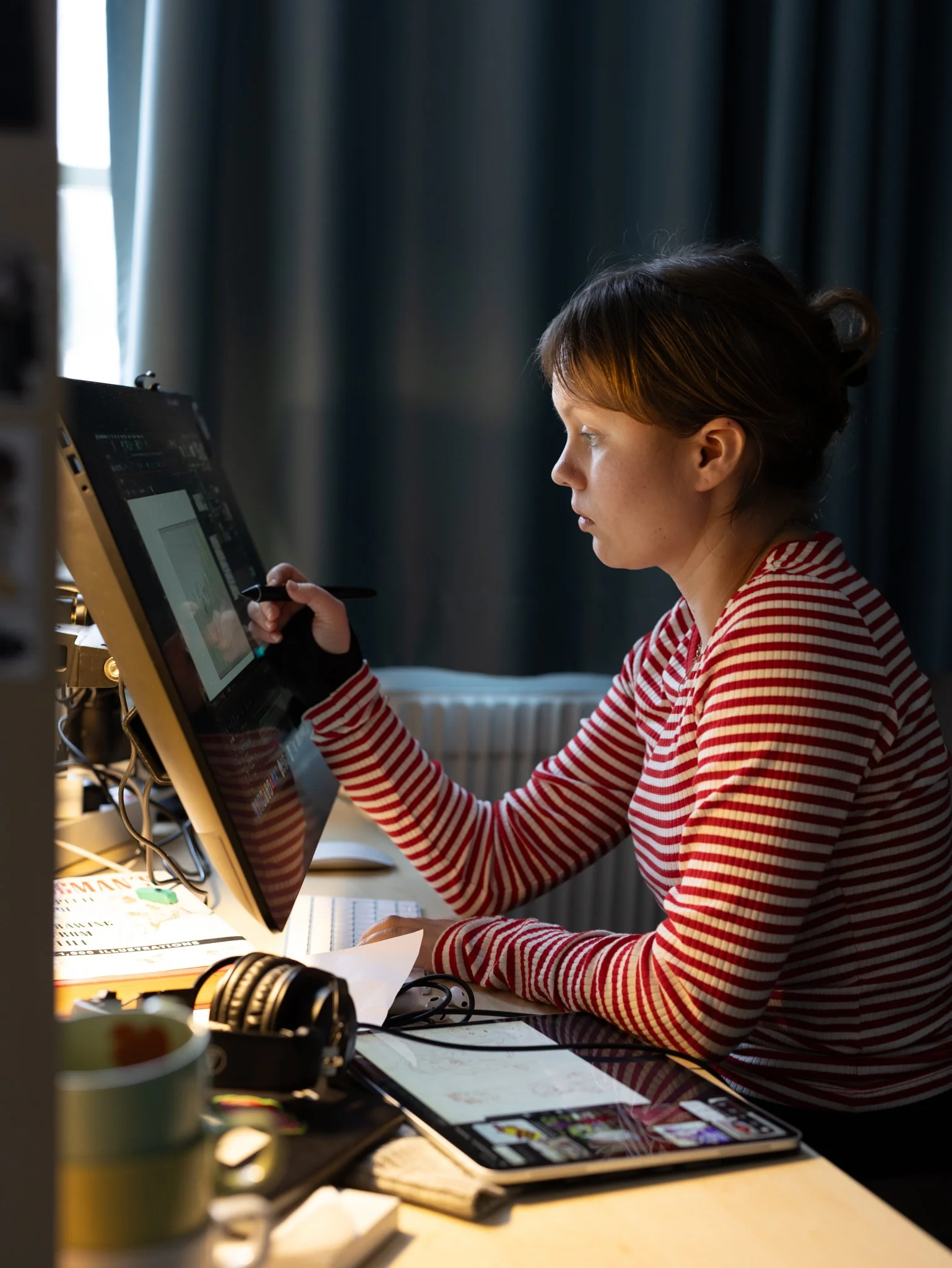 Kvinna med röd-vit randig tröja som arbetar vid en dator med ett penna i handen, på ett skrivbord med papper och elektronik. Bakgrund av mörka gardiner och fönster.