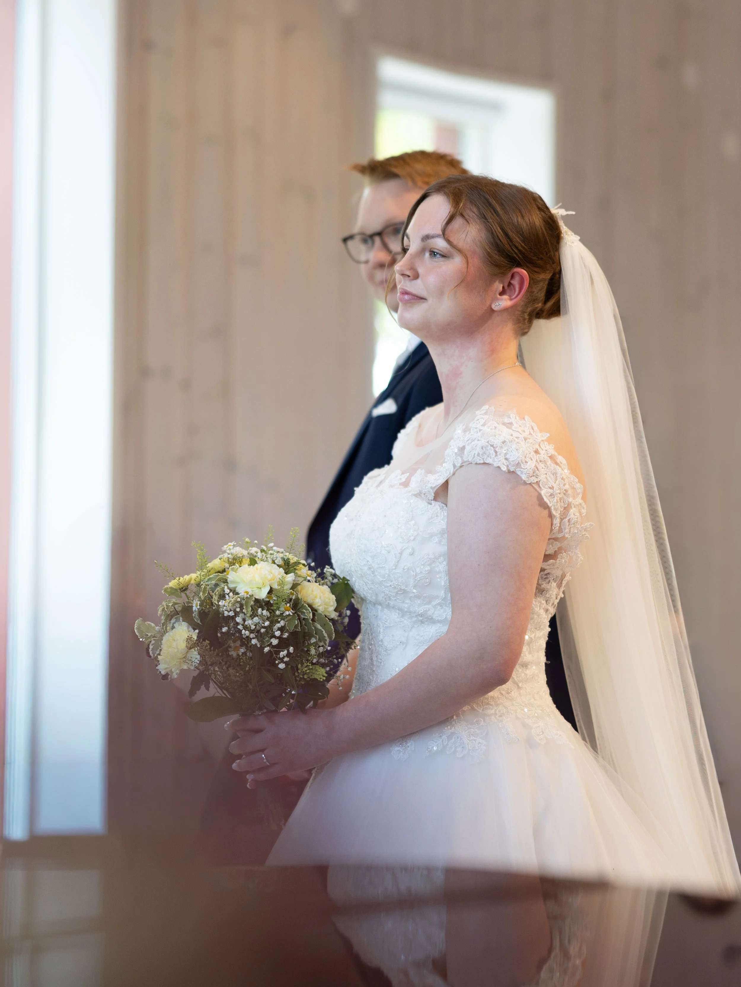 En brud har på sig en vit brudklänning och håller en bukett med gula och vita blommor. Hon har långt hår, delvis lockigt, och bär en slöja. Bakom henne står en man i mörk kostym och glasögon. De står i ett rum med träväggar och fönster.