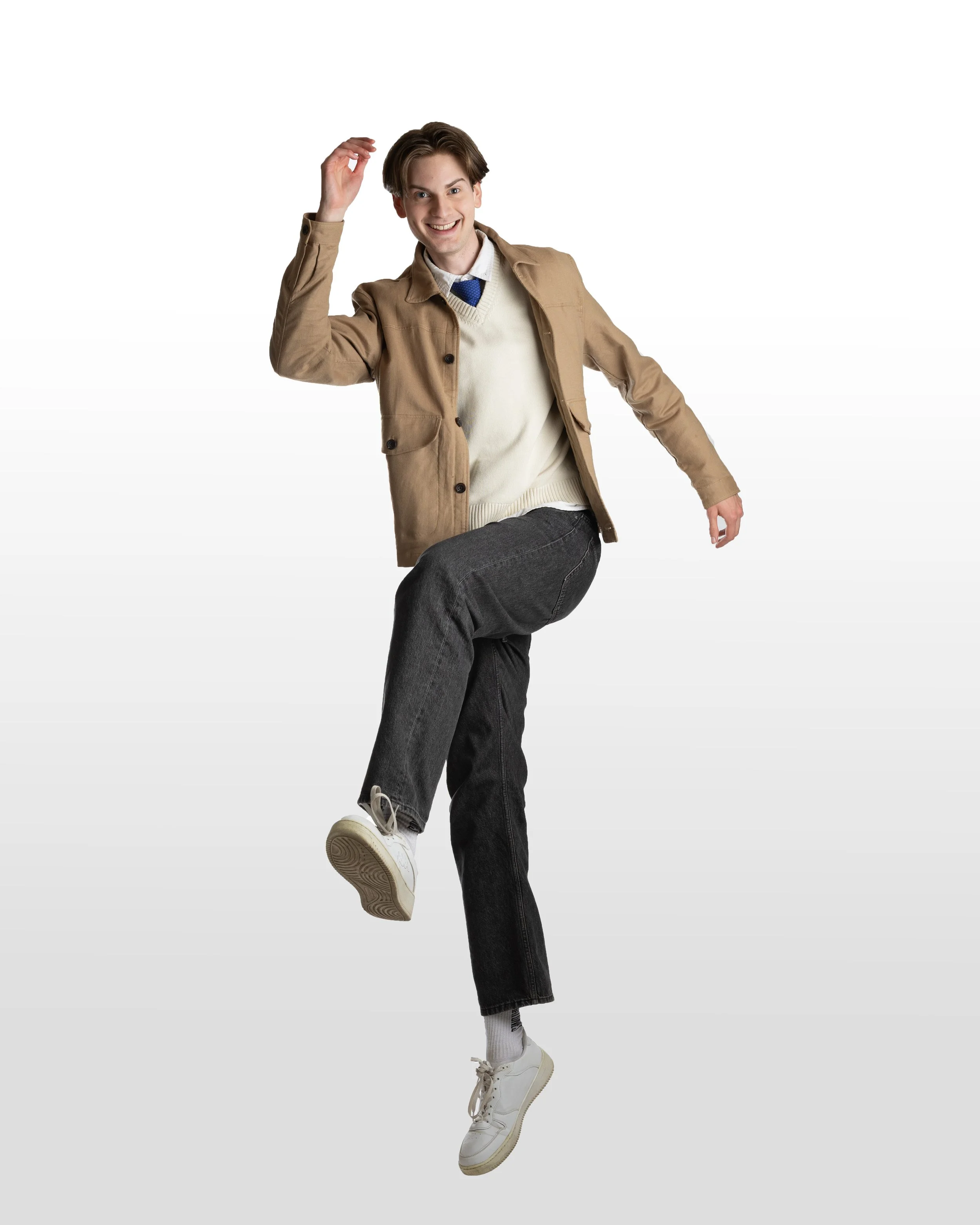 Ung man som dansar eller leker, klädd i beige jacka, vit kavaj, blå slipover, gråa byxor och vita sneakers, mot en vit bakgrund.