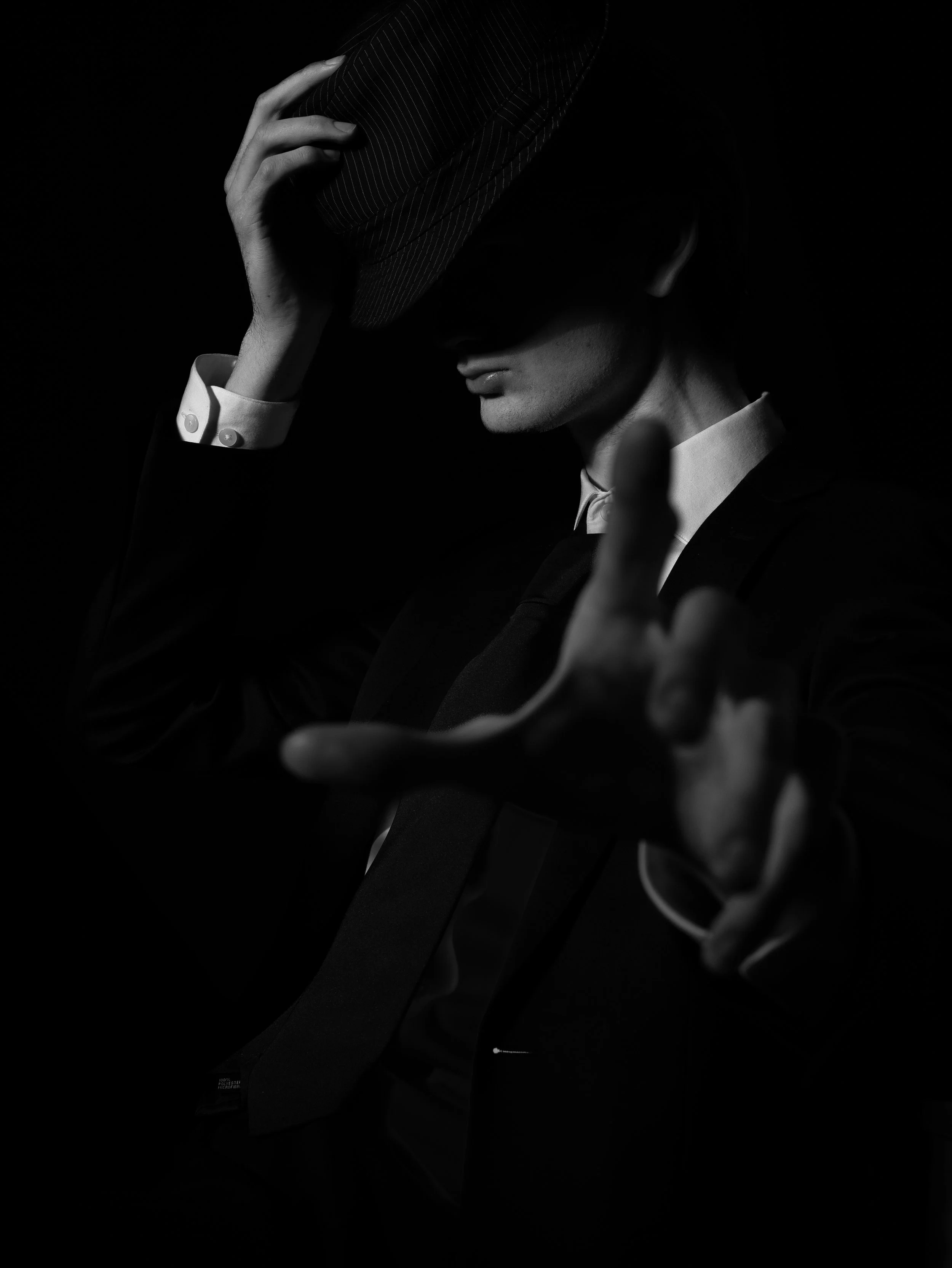 Svartvitt fotografi av en person som bär en mörk kostym och vit skjorta, håller en randig hatt i handen i mörkt ljus.