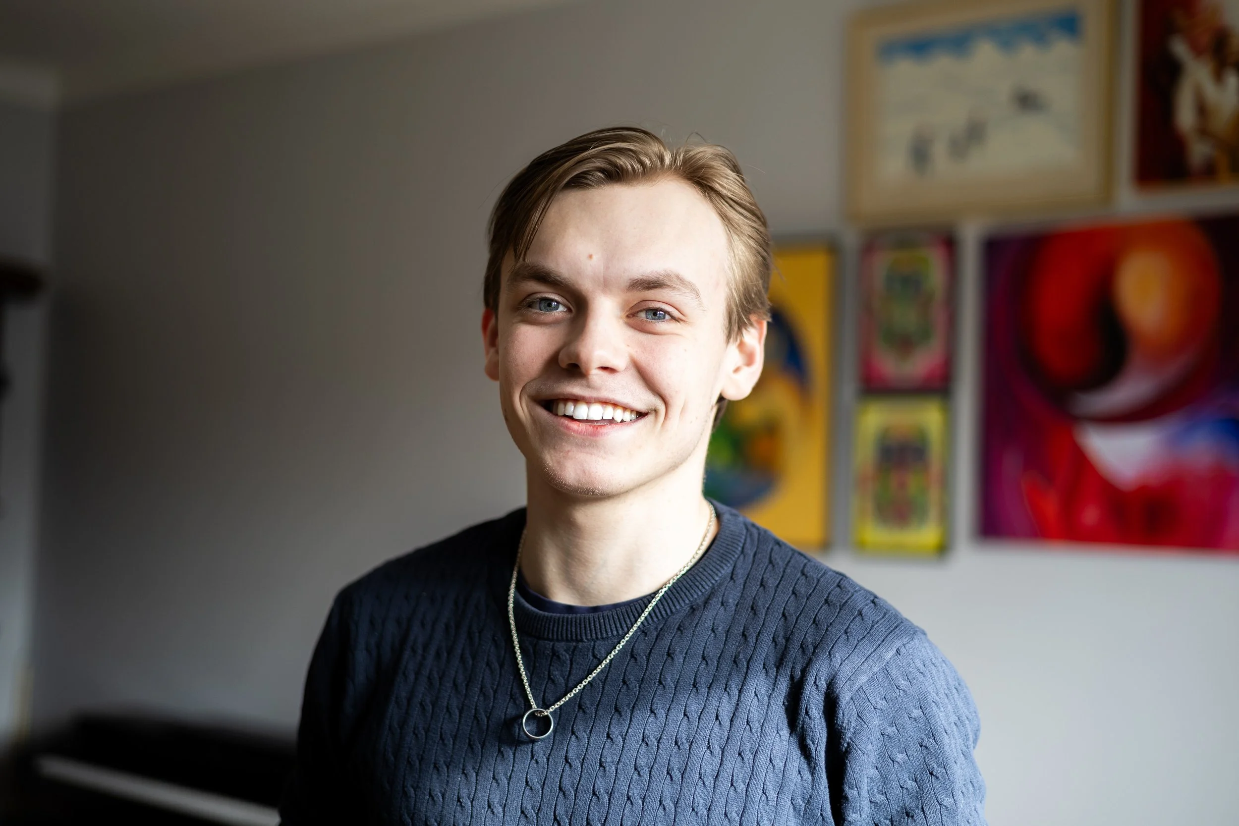 Ung man med ljus hud och blont hår, tänd ett leende, bär en mörkblå stickad tröja och halsband, står framför färgglada konstverk på en vägg.
