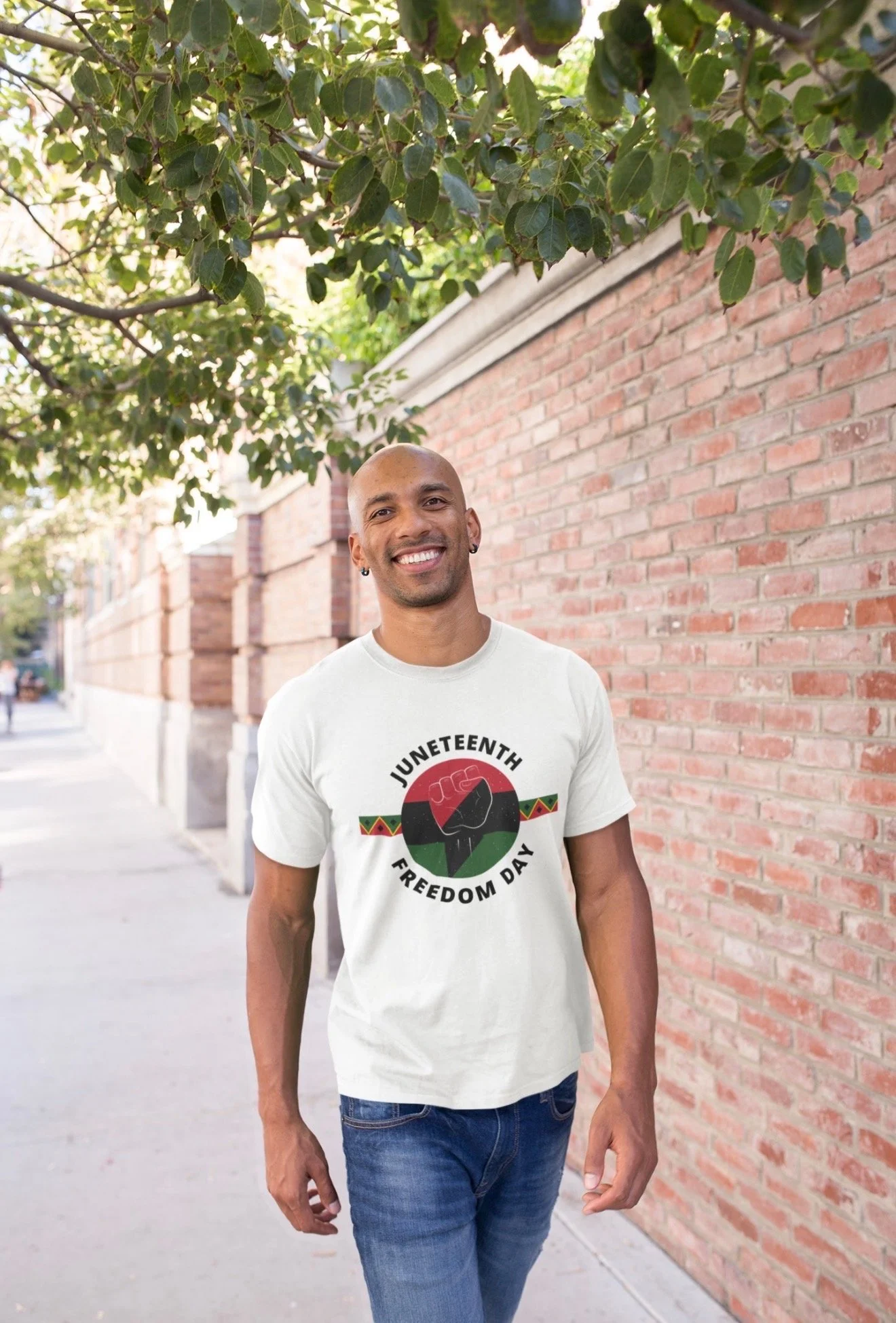 A smiling man with a shaved head and earrings, walking outdoors next to a brick wall and green tree leaves, wearing a white T-shirt with Juneteenth Freedom Day printed on it and blue jeans.