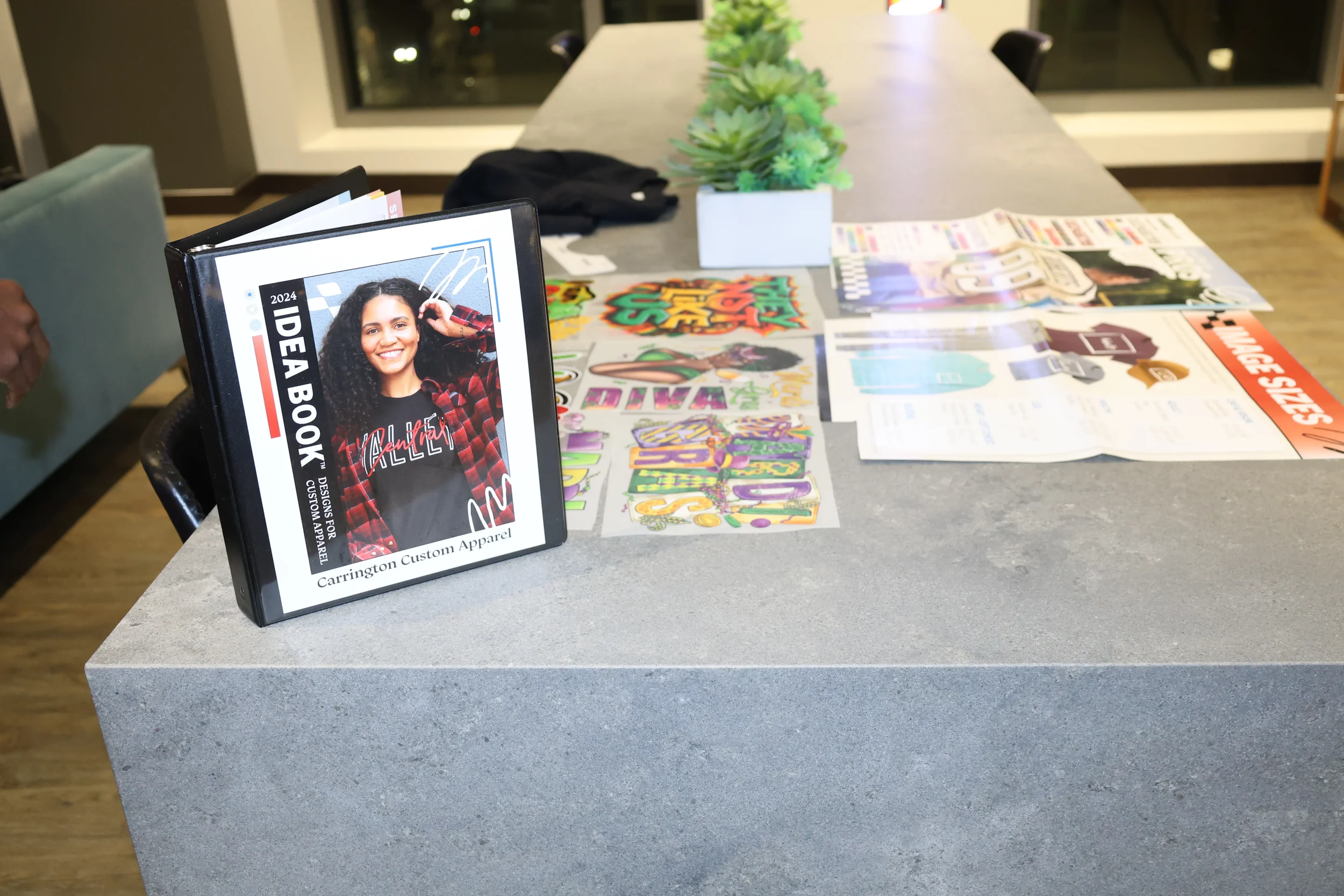 Table displaying various colorful magazines and pamphlets, with a small potted succulent plant and a framed photo featuring a smiling woman, at an indoor setting with large windows in the background.