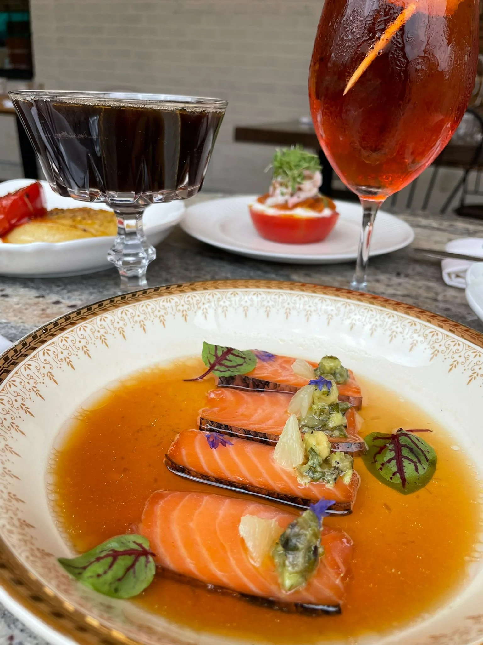 Une assiette avec du saumon, une sauce orange, des herbes et des fleurs comestibles. En arrière-plan, un verre de vin rouge, un verre de café et une tomate creusée avec une garniture.