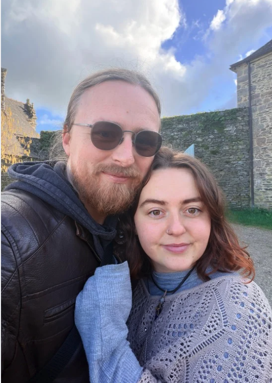 Deux personnes prennent un selfie devant un vieux mur en pierre avec un ciel partiellement nuageux.