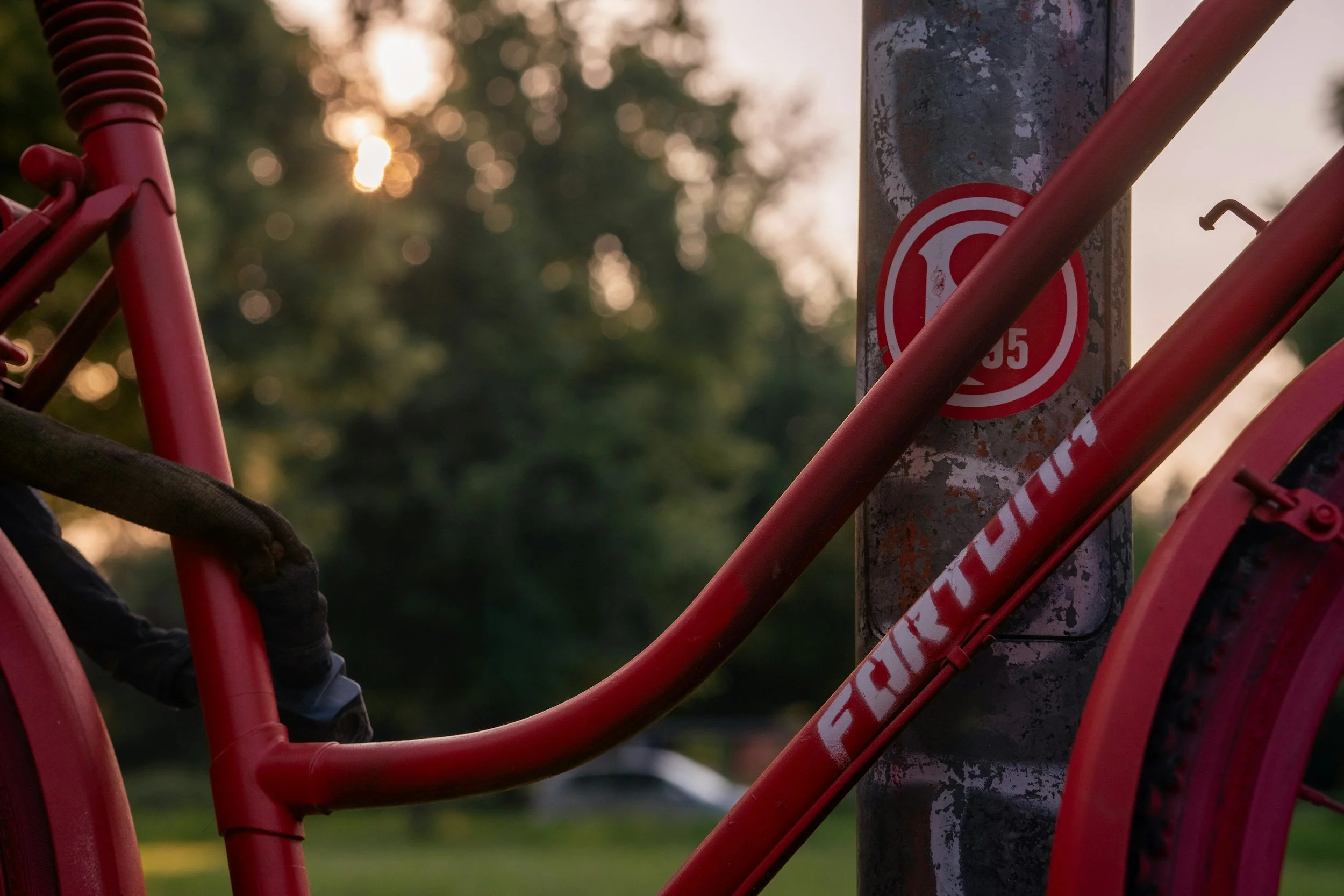 Rote Fahrradrahmen, Sattel und Fahrradsteig an einem Fahrrad, das an einem Metallpfosten befestigt ist, im Hintergrund Bäume bei Sonnenuntergang.