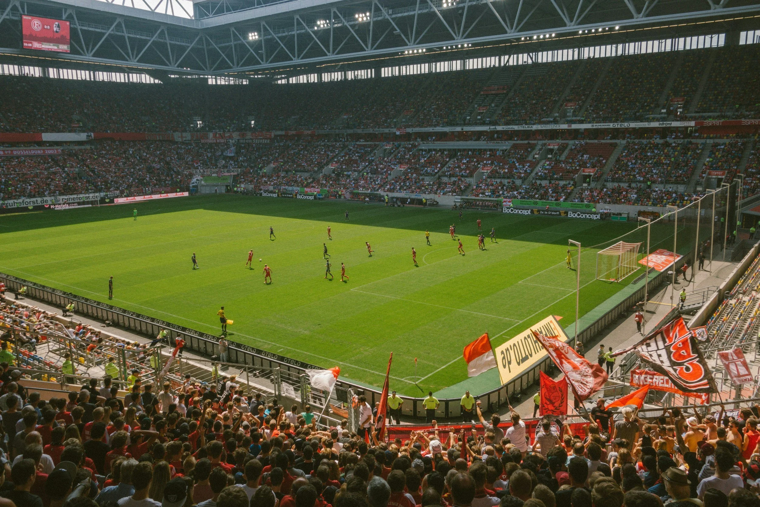 Fußballspiel im ausverkauften Stadion, Fans mit Fahnen und Trikots sitzen im Publikum, Spieler auf dem Feld, einige in roten Trikots, andere in dunklen.