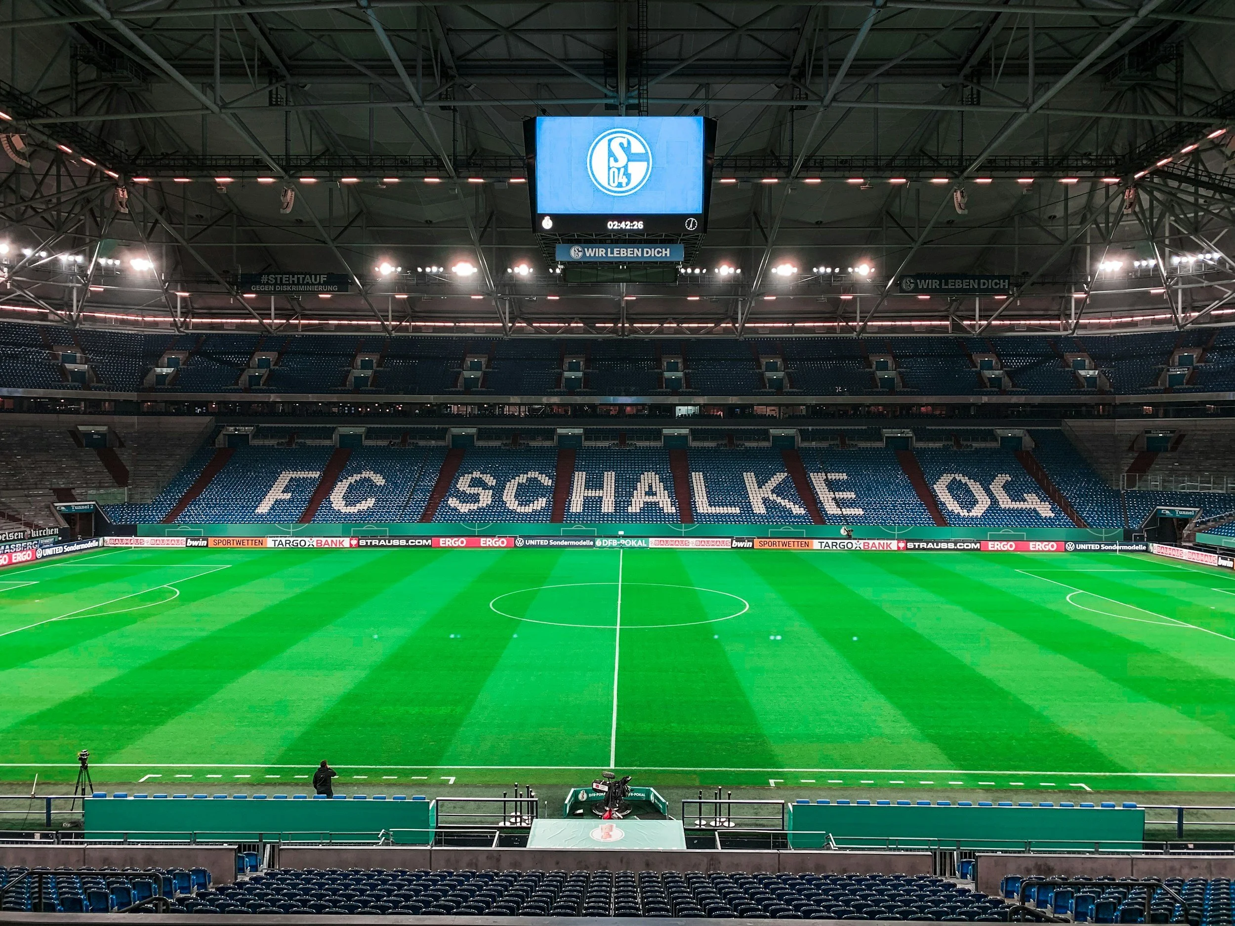 Leeres Fußballstadion mit grüner Spielfläche, leeren blauen und roten Sitzplätzen, einem großen Bildschirm mit Logo des FC Schalke 04, im Hintergrund eine Stadionrückwand mit Bannern und Lichtern.