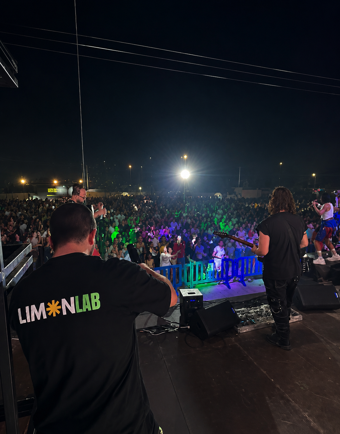 Concierto al aire libre por la noche con un público numeroso, músicos en el escenario y luces de colores.