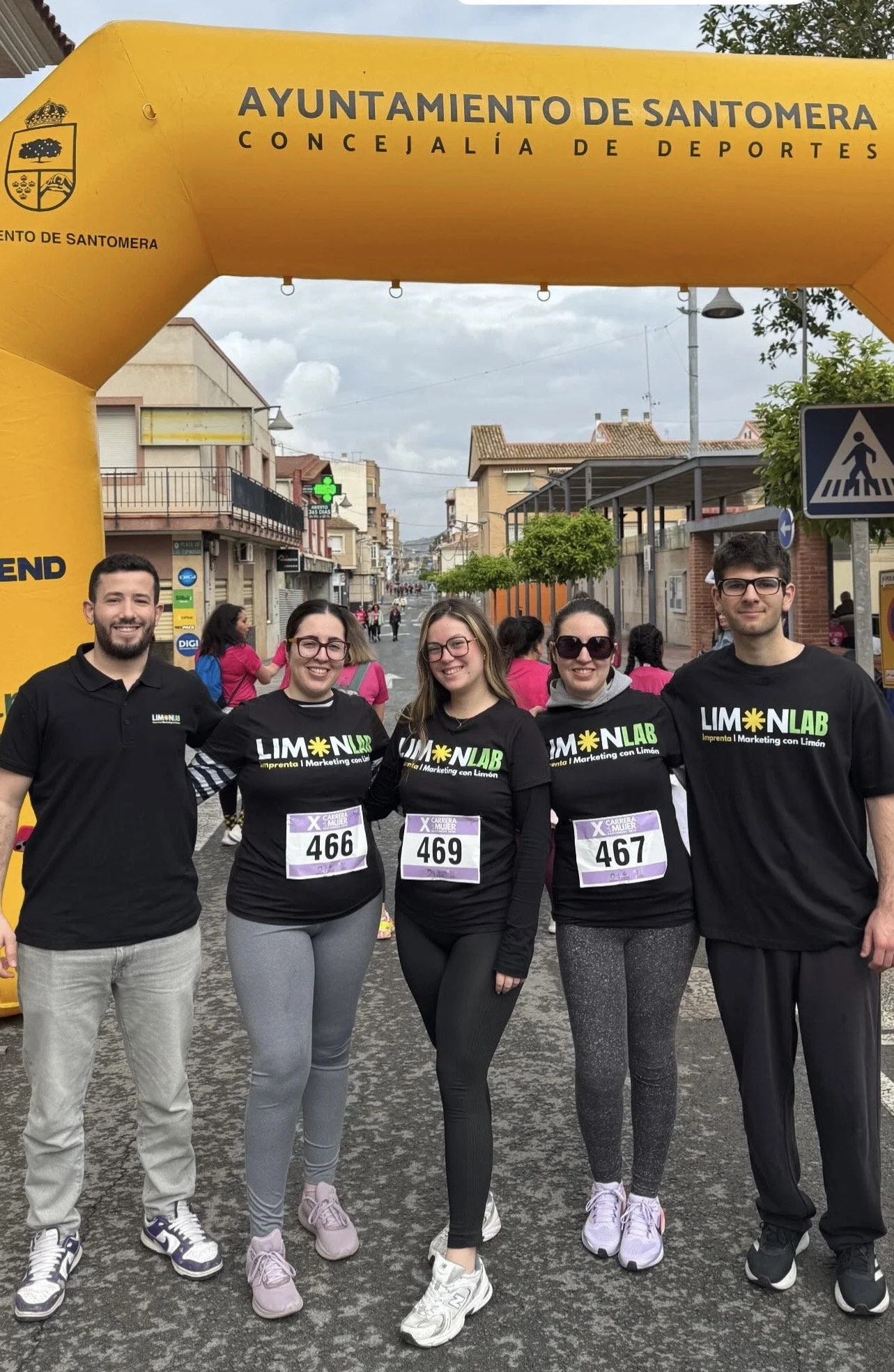 Cinco personas con ropa deportiva y números en la camiseta participan en una carrera, con una estructura amarilla que dice "Ayuntamiento de Santomera Calleja de Deportes" y un grupo de personas en el fondo.