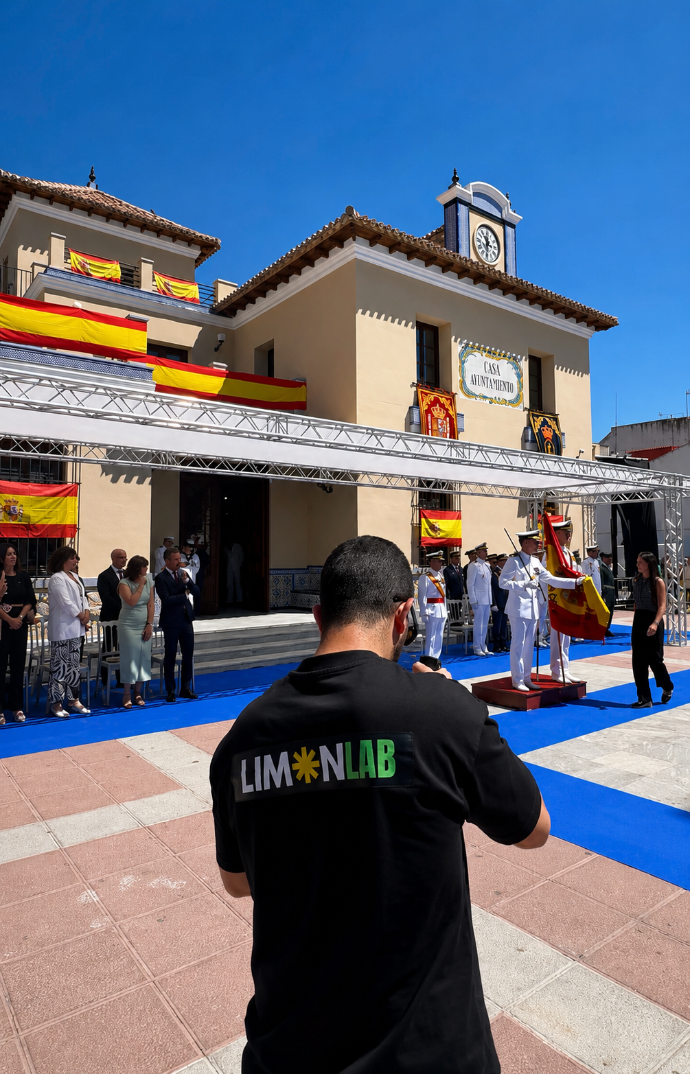 Ceremonia en la plaza del ayuntamiento decorada con banderas de España, donde oficiales en uniforme participan y varias personas asistiendo.