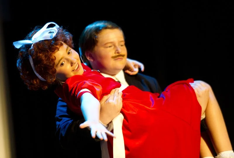 Two children performing on stage; one girl with curly hair, wearing a red dress and white bow, being held by a boy in a black jacket with a fake mustache.