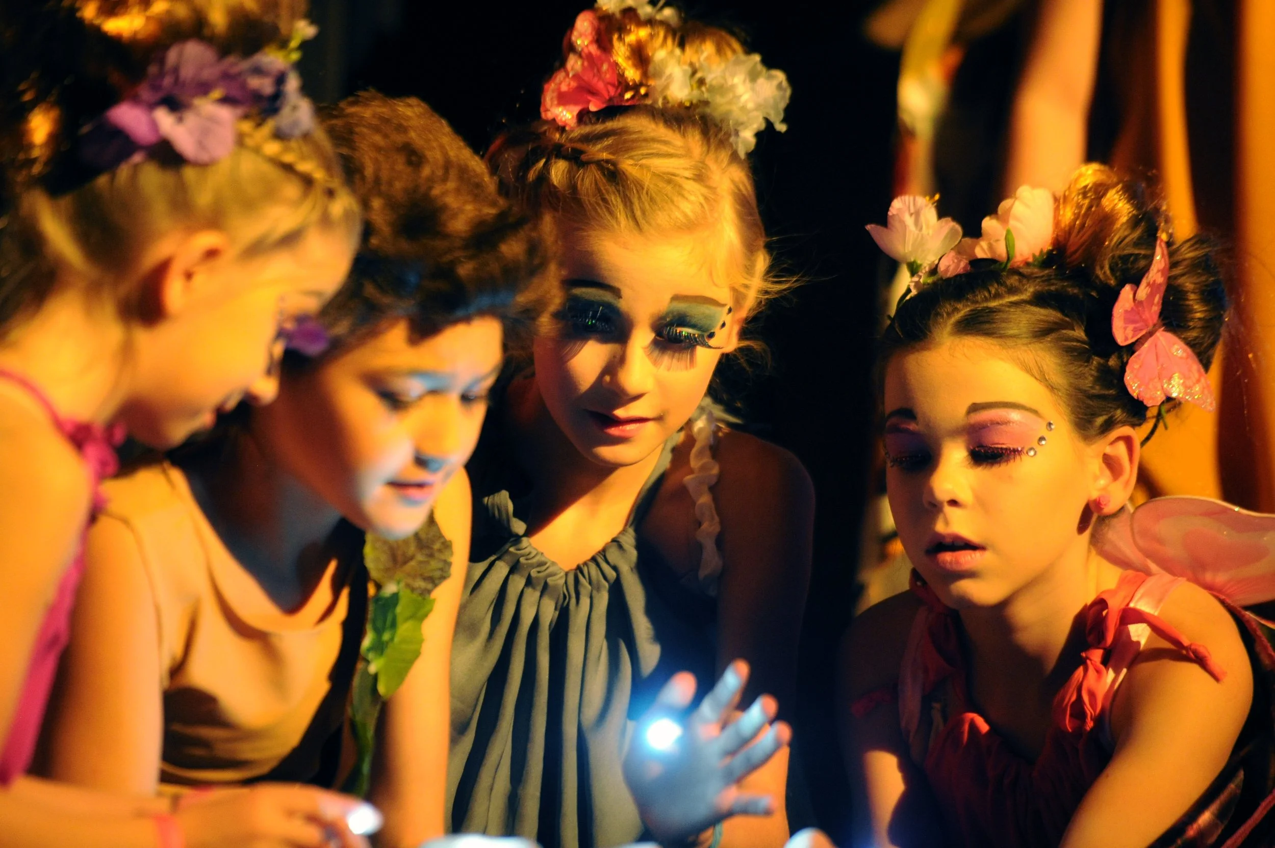 A group of young girls dressed in colorful festival costumes with floral headbands and makeup, gathered closely around a smartphone at night.