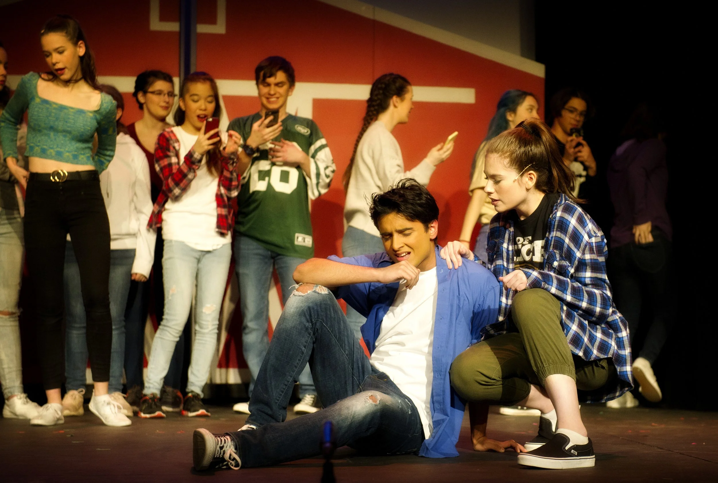 A group of young people on stage, with some taking photos or videos on their phones and others observing. Two individuals in the foreground are engaged in an activity, one sitting and the other kneeling on the floor.