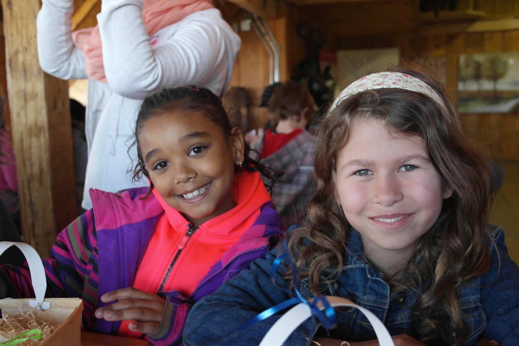 Smiling young girl with curly hair and light blue eyes, wearing a denim jacket, sitting at a table with another smiling girl with dark hair in a ponytail, wearing a colorful jacket and pink scarf, in a wooden room with other children in the backgroun