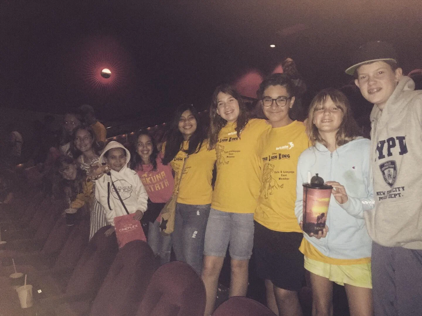 A group of children and teenagers is standing together in a dimly lit indoor space, smiling at the camera. Some are wearing yellow T-shirts with a lion graphic and text, and one is holding a drink. Others are wearing casual clothing and jackets, and 