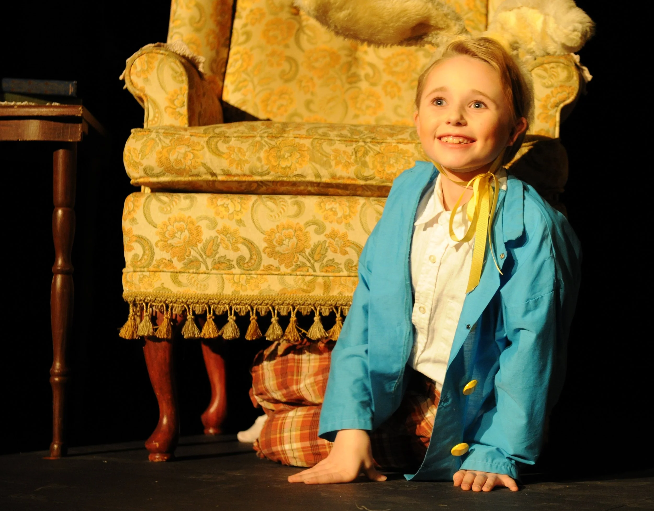 A young boy with light brown hair and blue eyes smiling while sitting on the floor with his hand on the ground. He is wearing a white shirt, a yellow tie, and a bright blue jacket. Behind him, there is a vintage yellow armchair with a floral pattern 