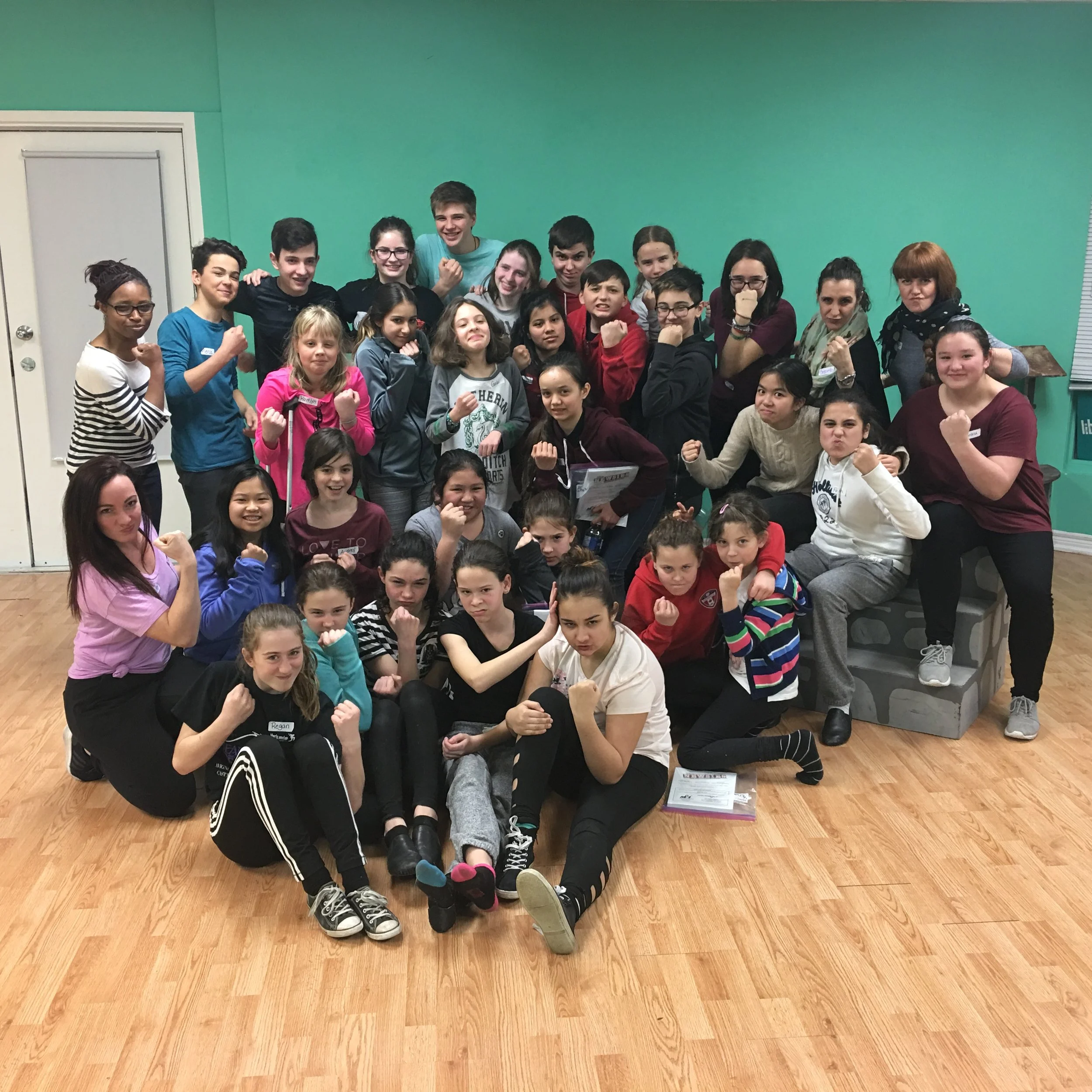 A large group of children and a few adults in a room with green walls, some children are sitting on the floor while others stand. Several people are making fists and their facial expressions show determination or excitement.