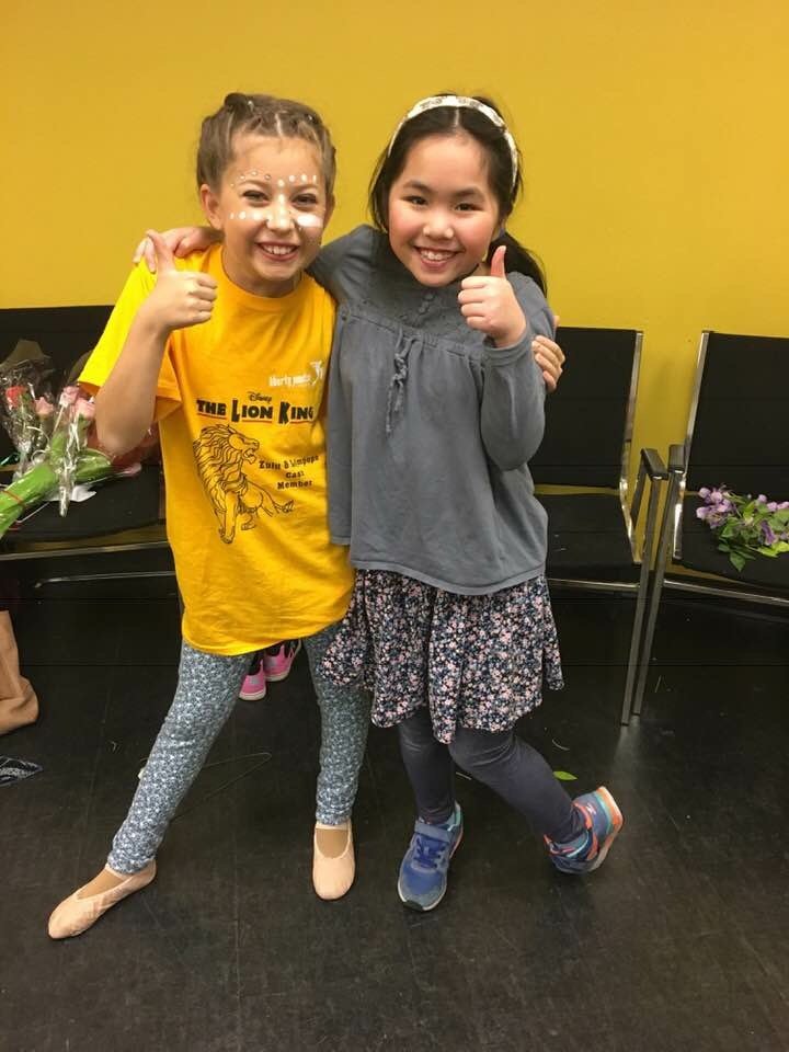Two young girls hugging and giving thumbs-up, smiling at the camera. One girl is wearing a yellow 'The Lion King' T-shirt and patterned leggings, the other girl is wearing a gray top with a floral skirt and sneakers. They are indoors against a yellow