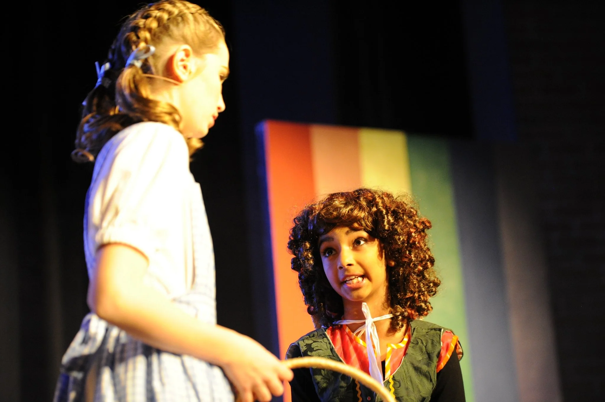 Two young girls with curly hair are on stage engaging in a conversation or performance, with a colorful background behind them.