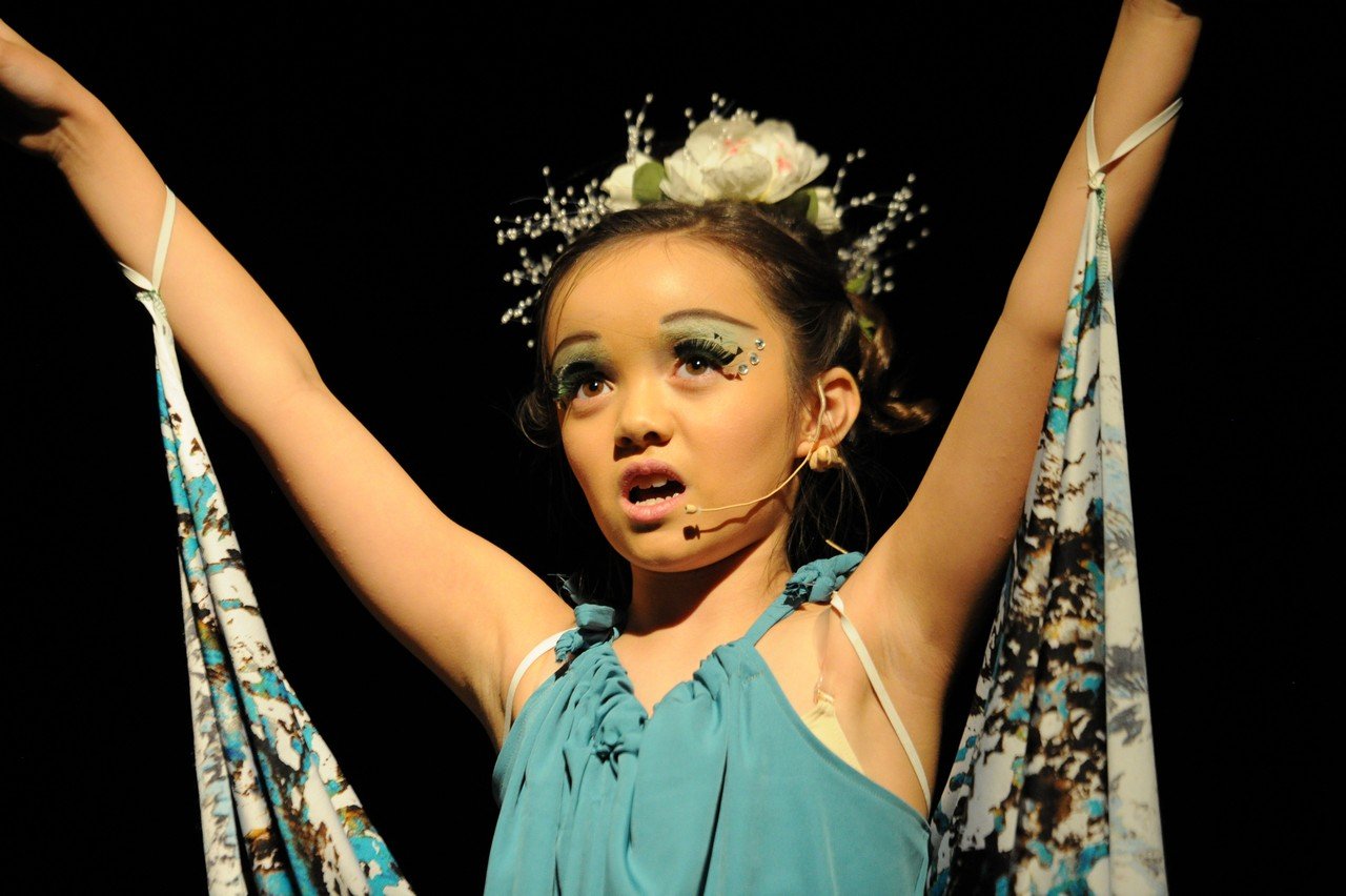 A young girl dressed in a blue costume with makeup and decorative elements performs on stage, with her arms raised, against a black background.