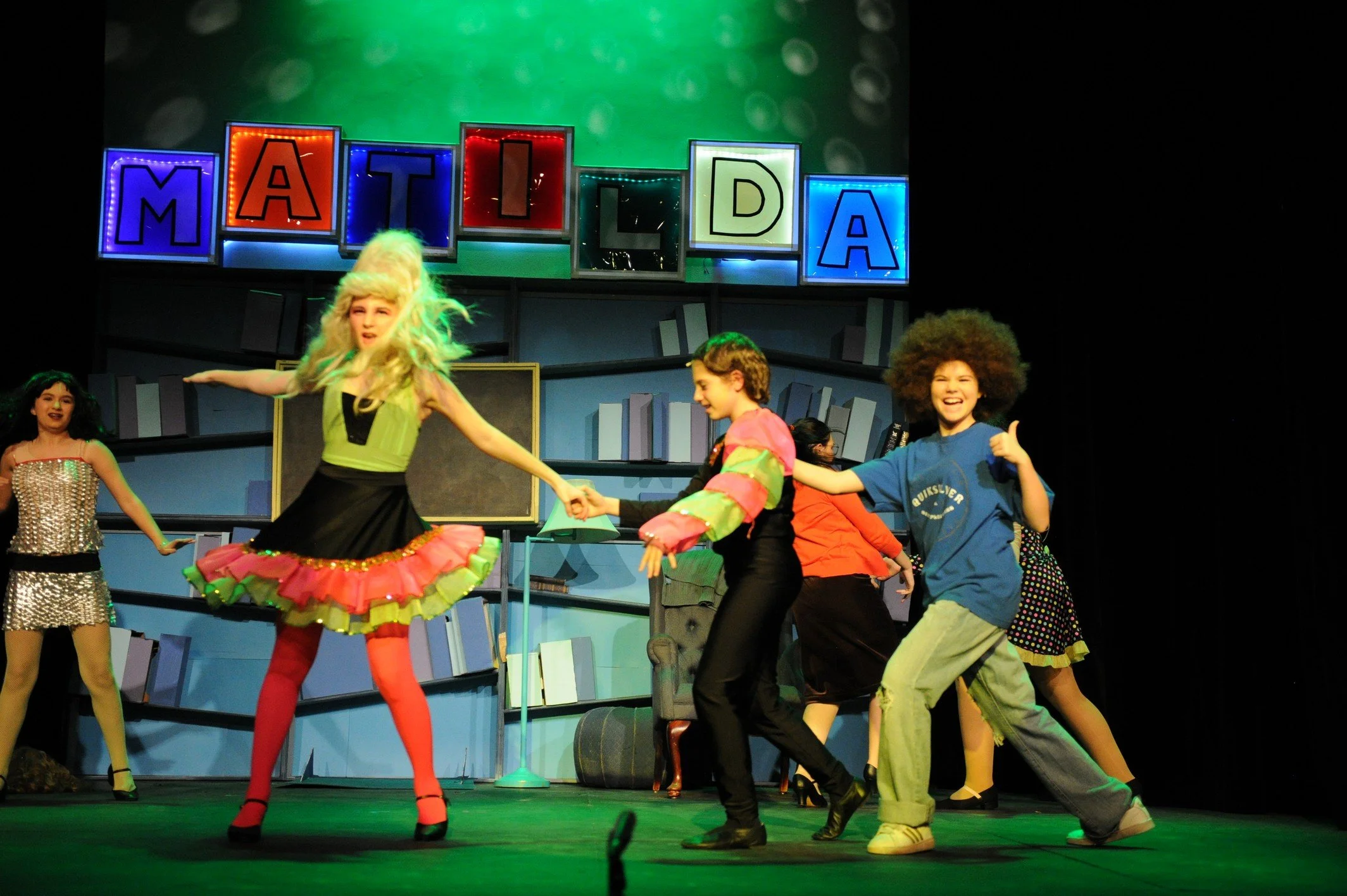 Children performing on stage with a background spelling 'MATILDA' in colorful lights.