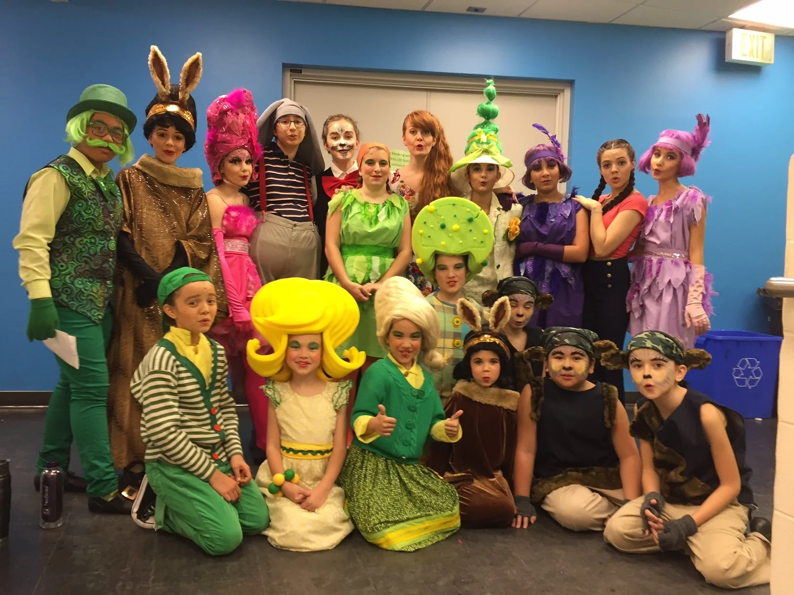 Group of children and adults in colorful costumes and makeup, posing for a photo backstage or at a costume event.