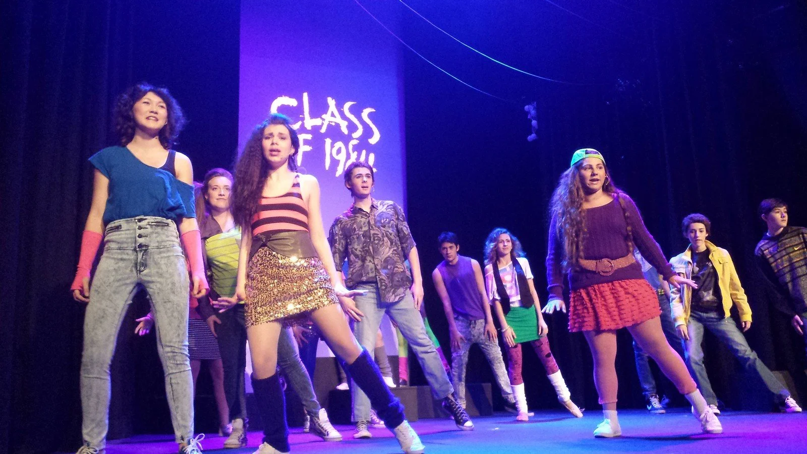 Group of teenagers performing on stage in costume, under purple lighting, with a sign that reads "Class of 1994" in the background.