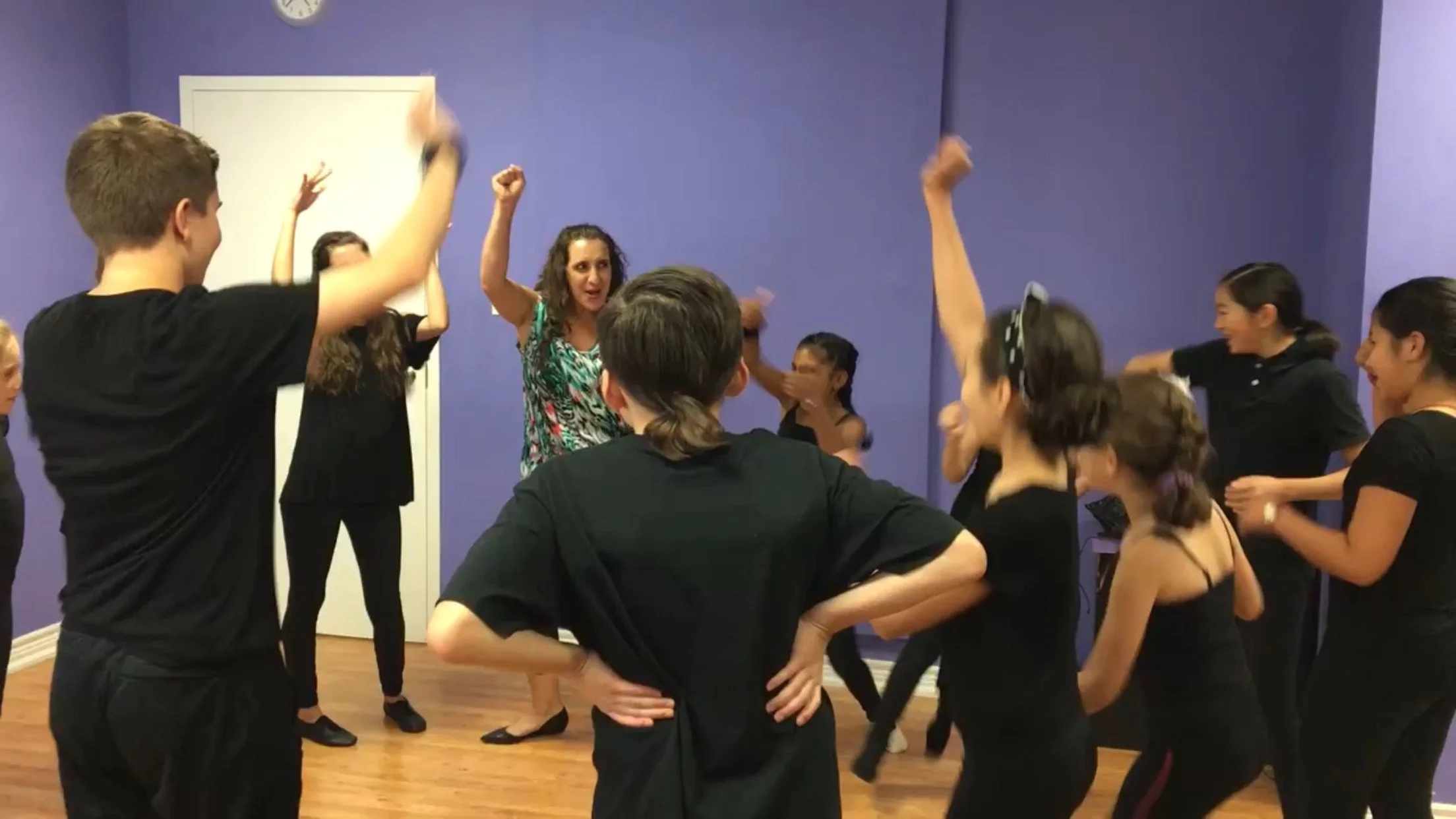 A group of people participating in a dance or fitness class in a room with purple walls. They are dressed in black and are moving energetically, with some raising their arms and others clapping or laughing.
