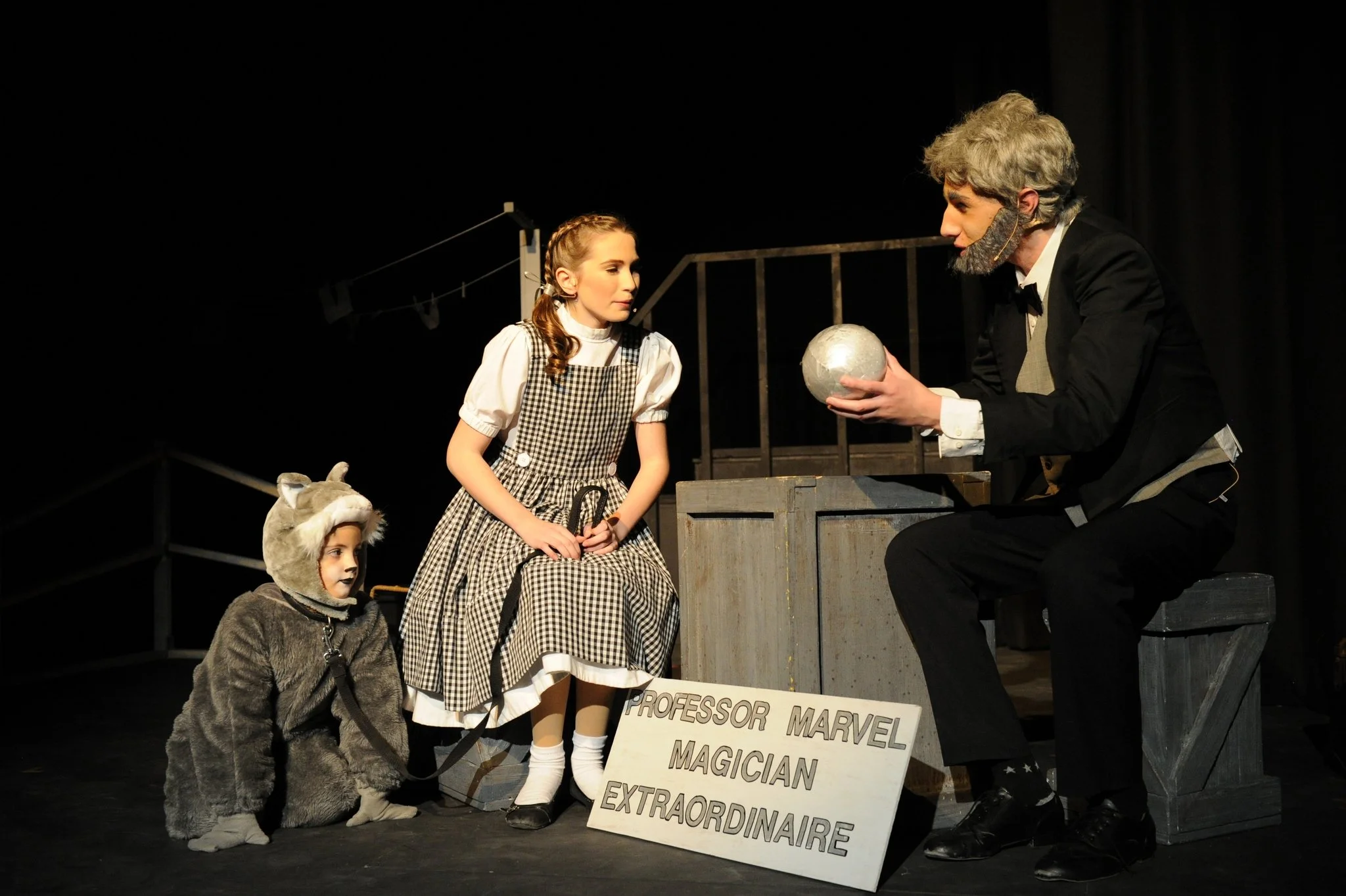 A theatrical stage performance featuring a man in a tuxedo with gray hair and a beard, holding a metallic globe, engaging with a girl in a checkered dress. Two children in animal costumes, one with a wolf costume and the other with a mouse costume, w