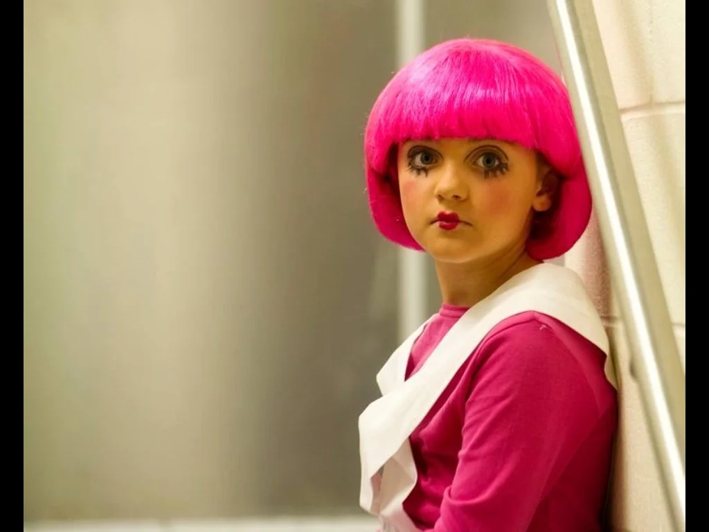 A young woman with bright pink hair styled in a bob, wearing a pink outfit with white accents, standing against a wall and looking directly at the camera.