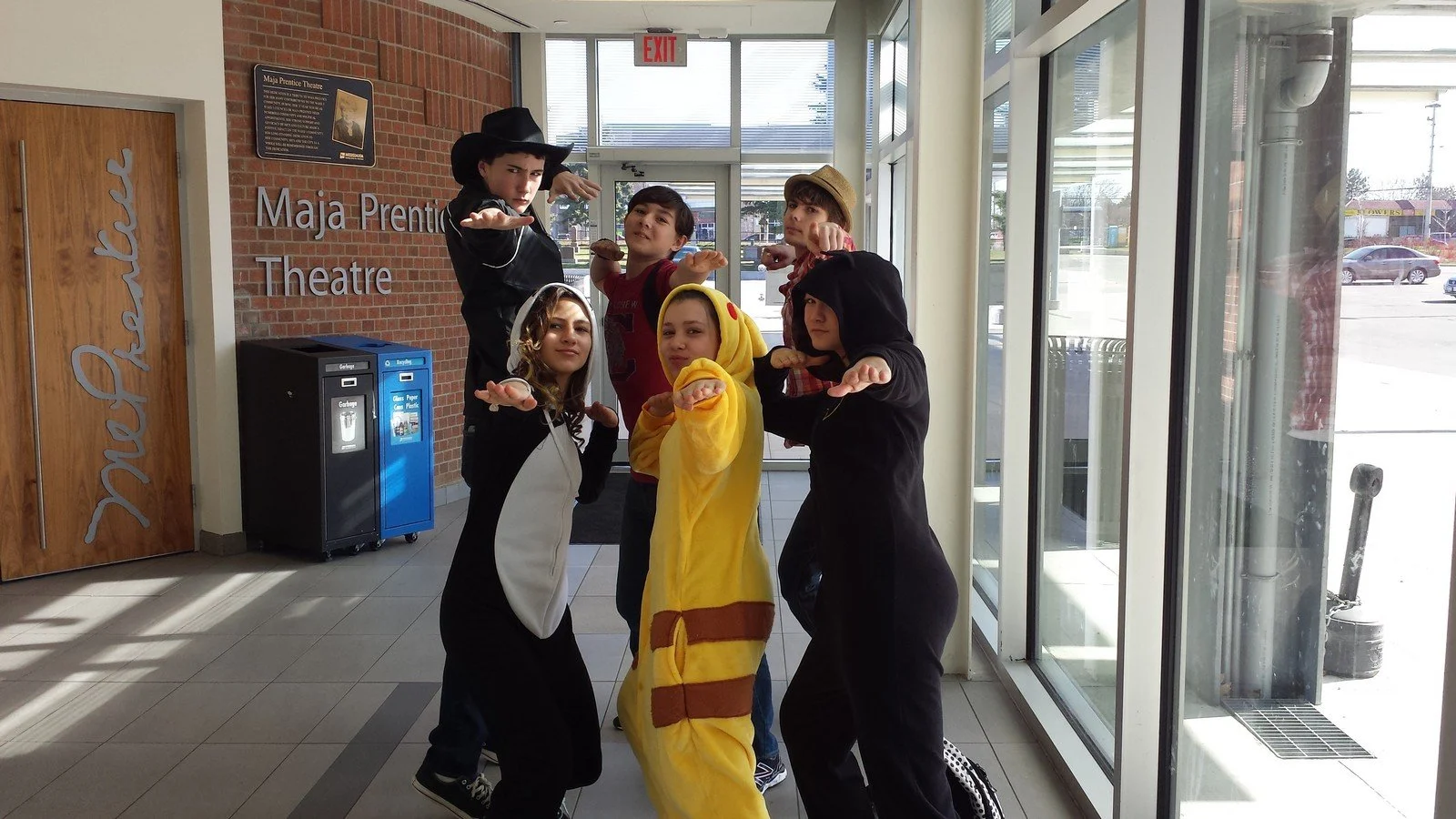 A group of six children dressed in various costumes, including a witch, a lion, and a hoodie, posing in a theater lobby with glass doors, brick wall, and a sign that reads 'Maja Prentice Theatre' in the background.
